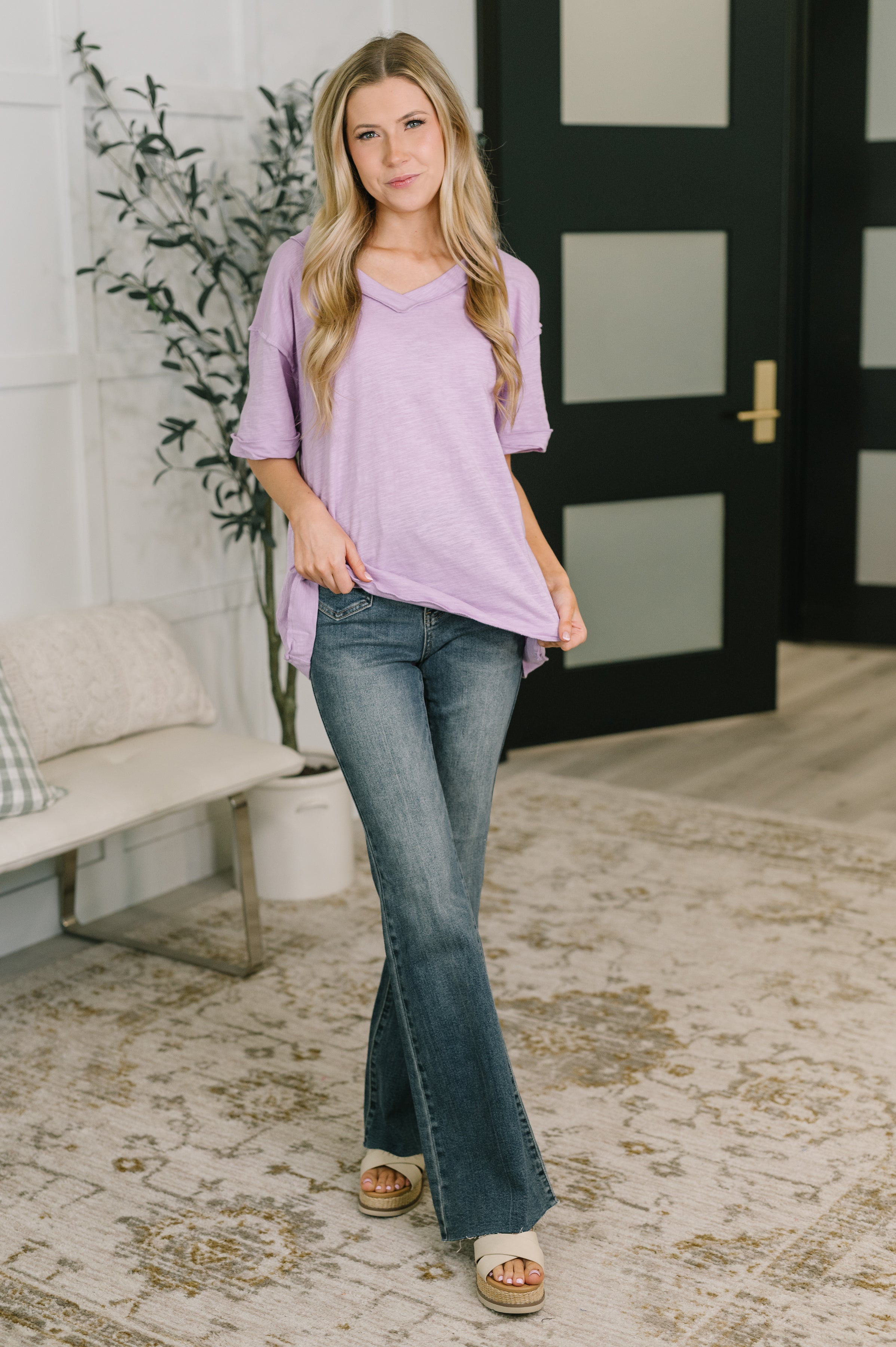 Woman wearing lilac relaxed V-neck tee paired with flared jeans and sandals in bright modern room with rug and greenery