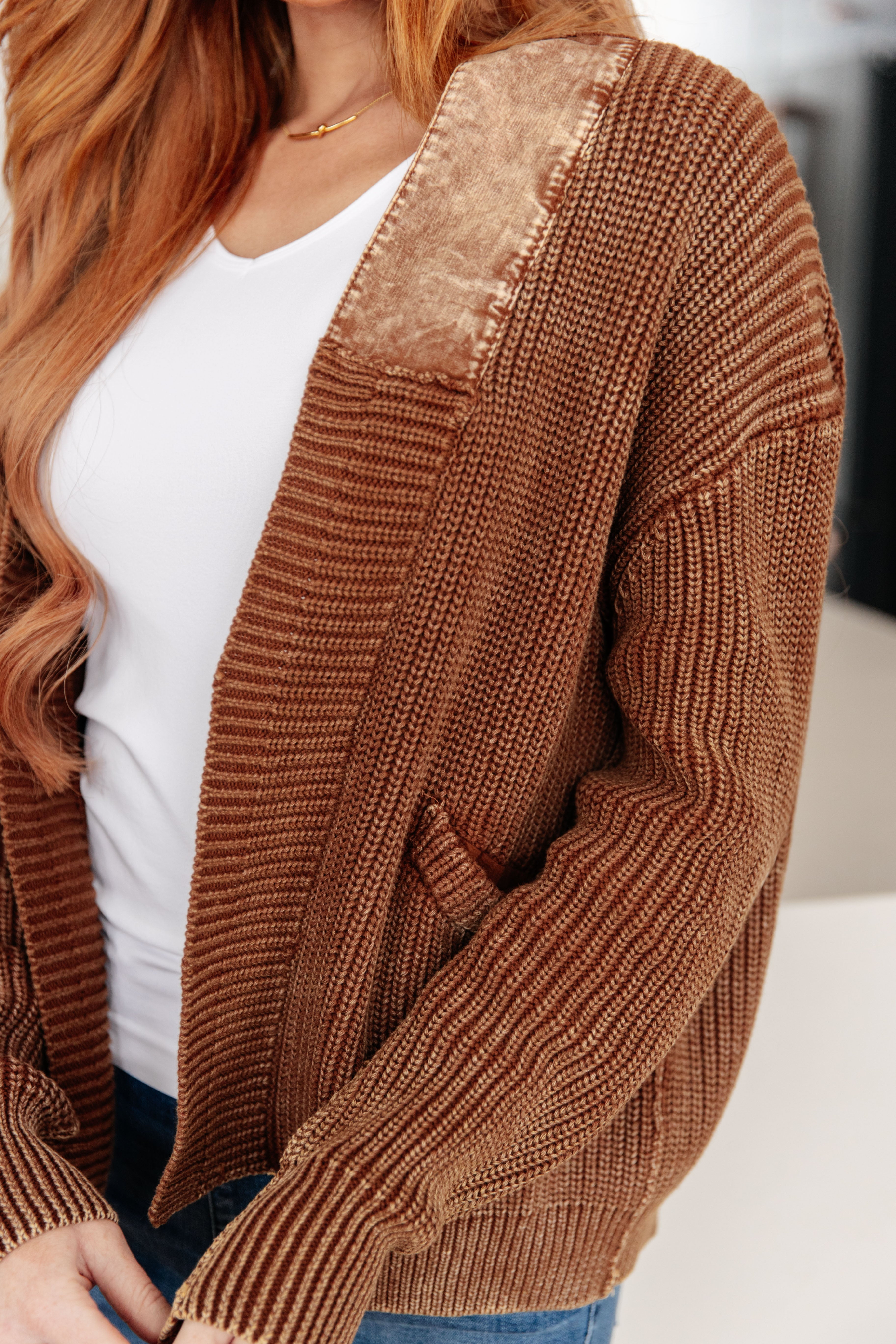 Close-up of a brown knit cardigan with velvet collar detail worn over a white top and blue jeans indoors