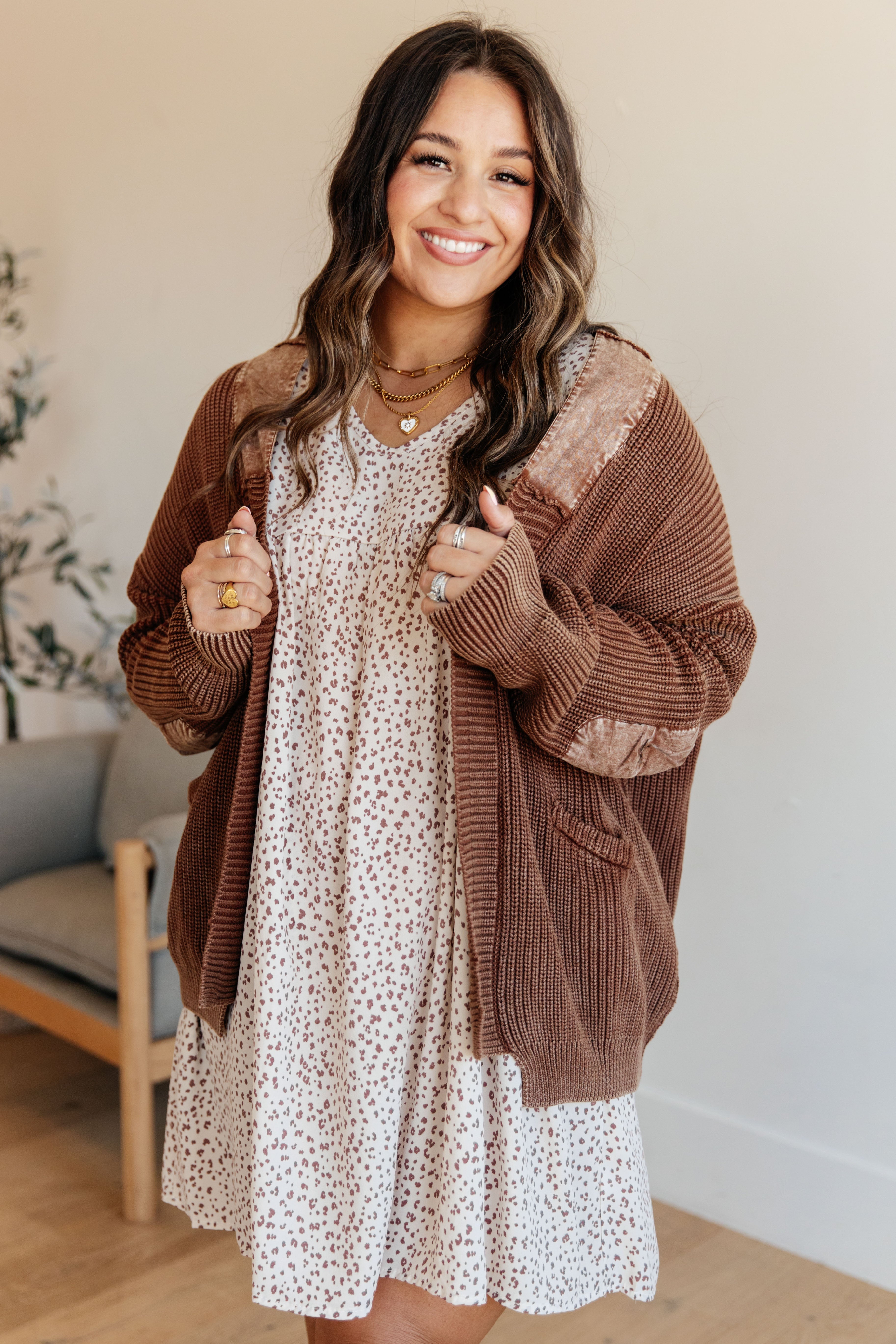 Smiling woman wearing a brown knit cardigan with velvet patches over a white patterned dress in bright indoor setting