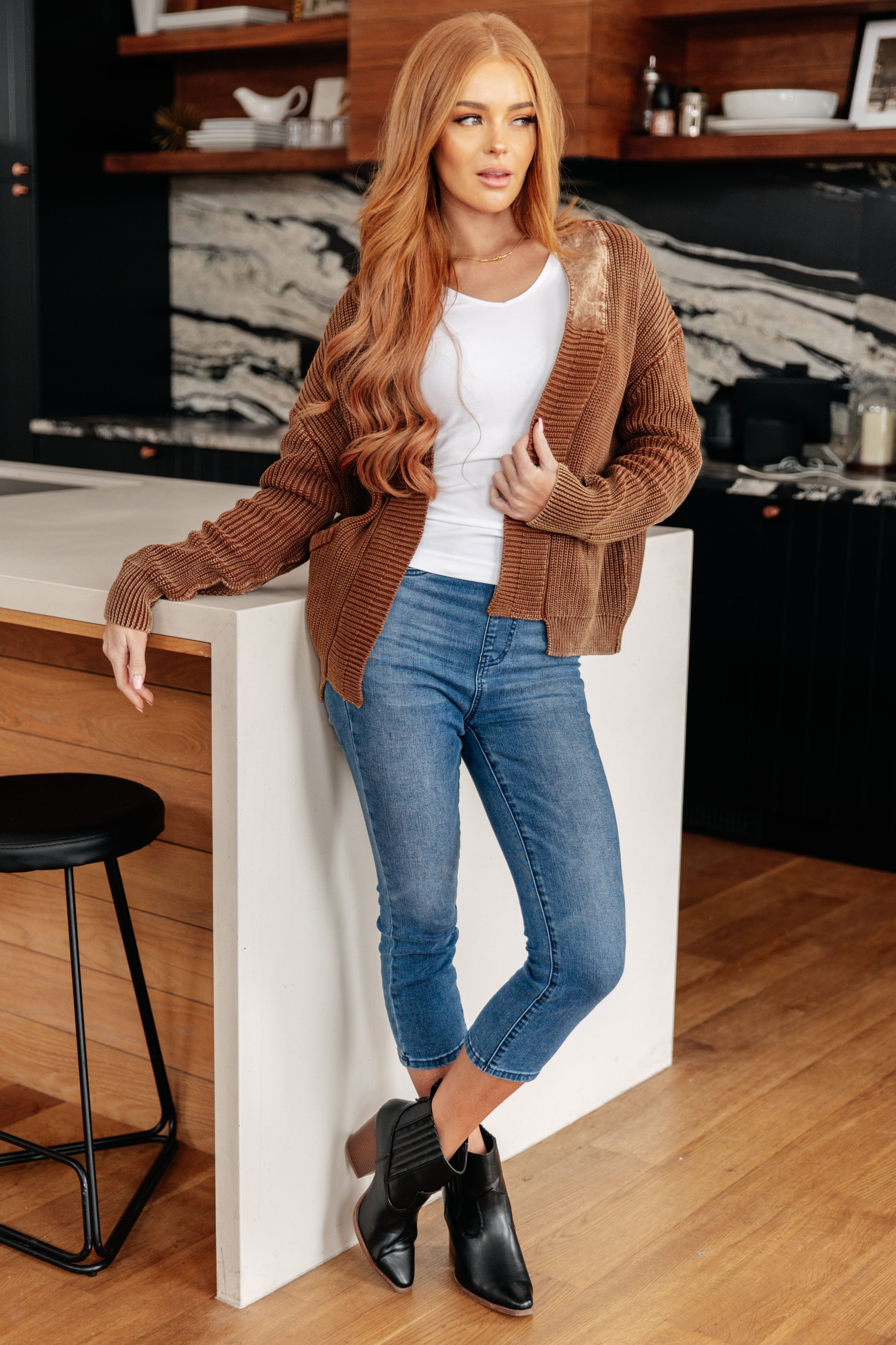 Model wearing a brown plush cardigan over a white top with cropped blue jeans and black ankle boots indoors