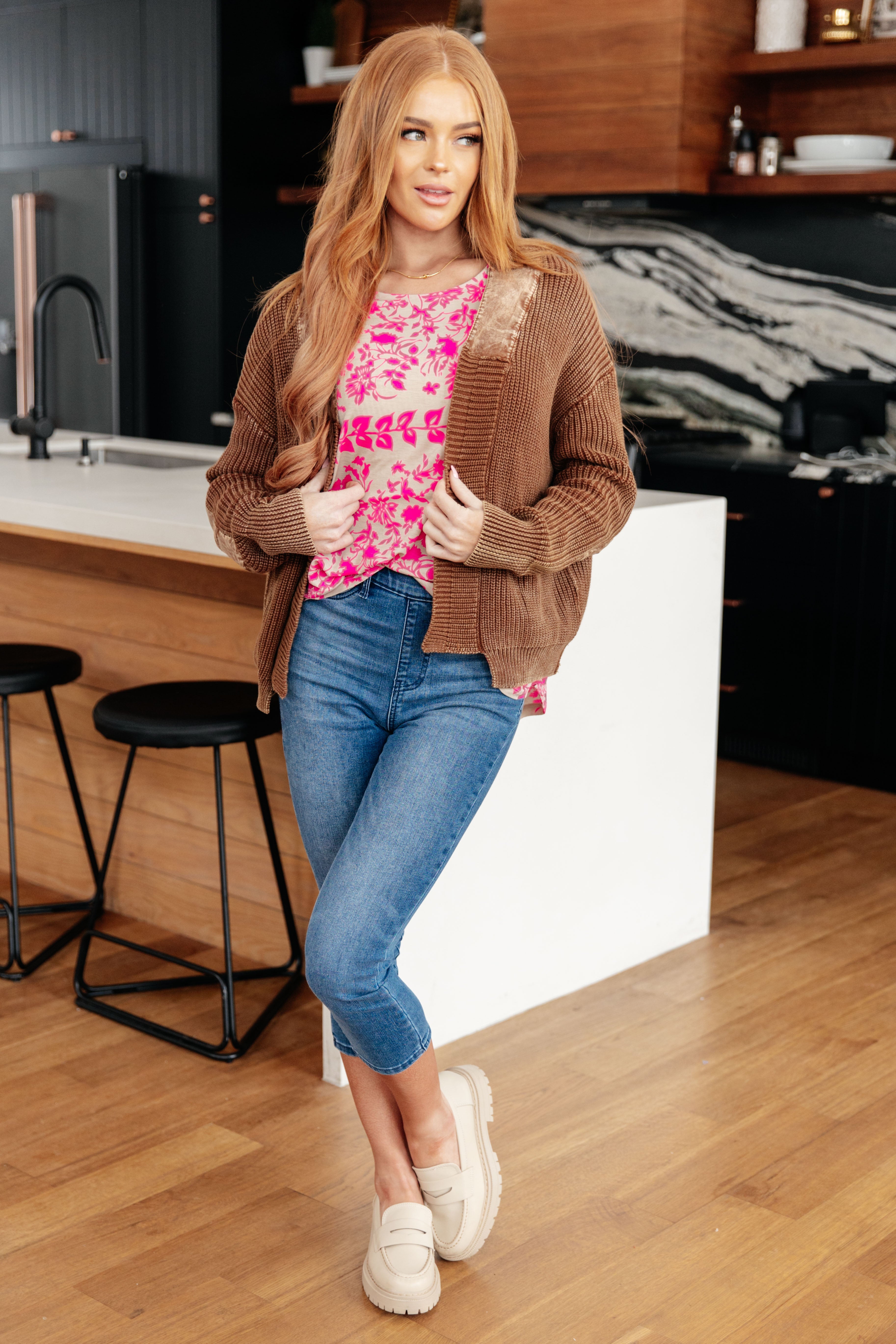 Woman wearing a brown cozy cardigan over a pink floral top paired with cropped blue jeans and cream shoes indoors