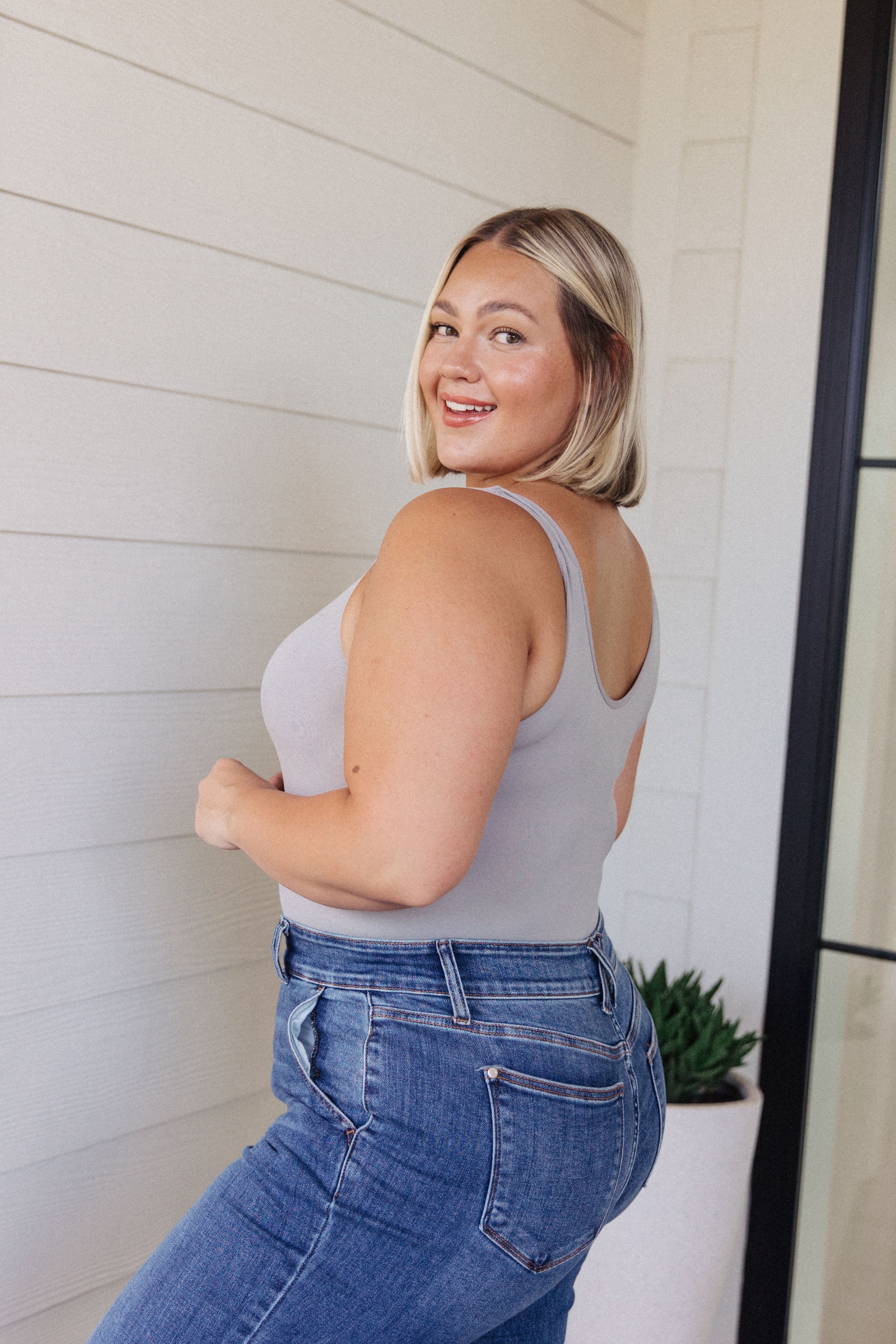 Smiling woman with blonde hair wearing a grey sleeveless fitted top and high-rise blue jeans standing indoors