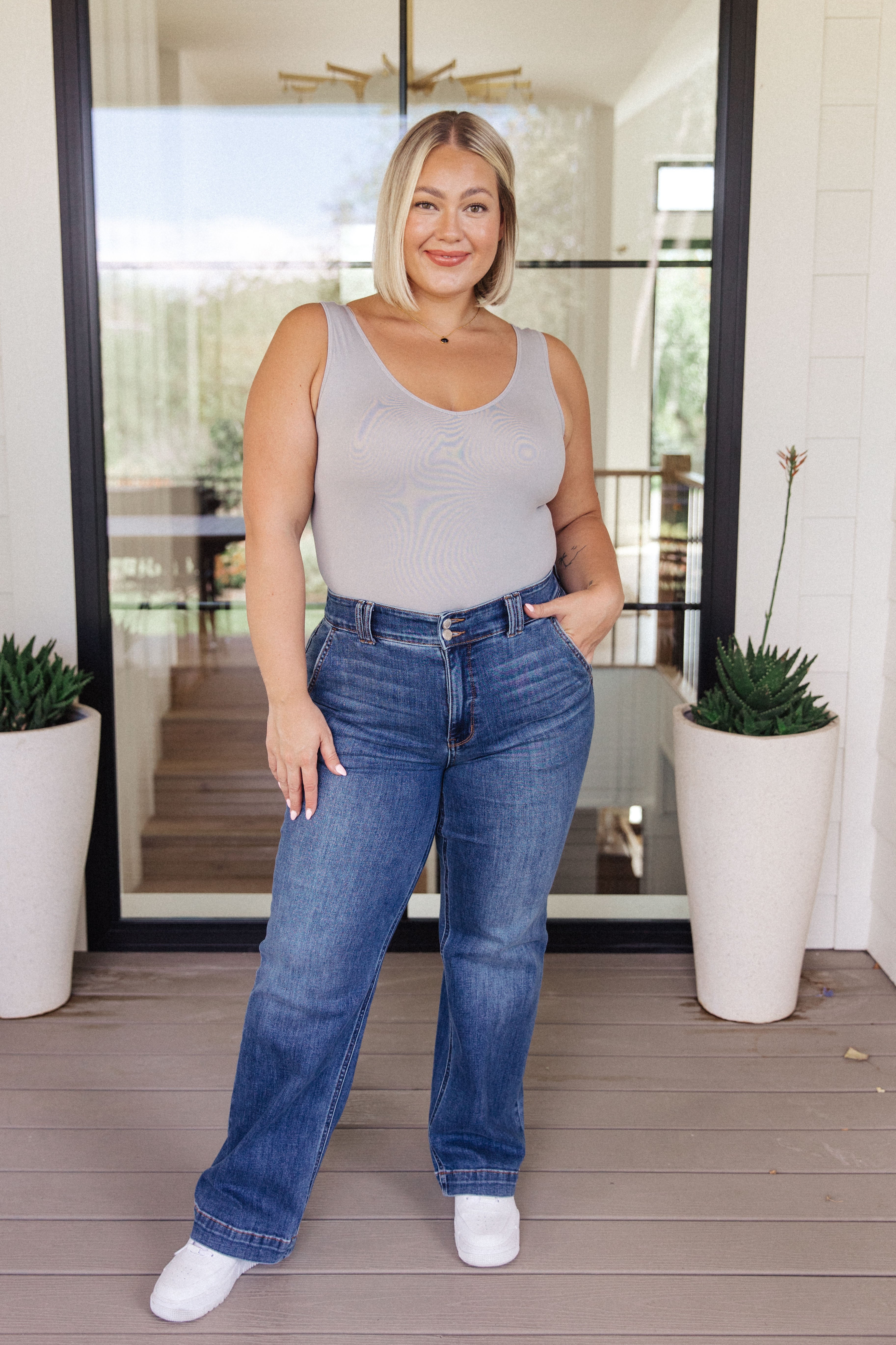 Confident woman with blonde bob wearing a grey sleeveless bodysuit tucked into relaxed-fit blue jeans standing indoors