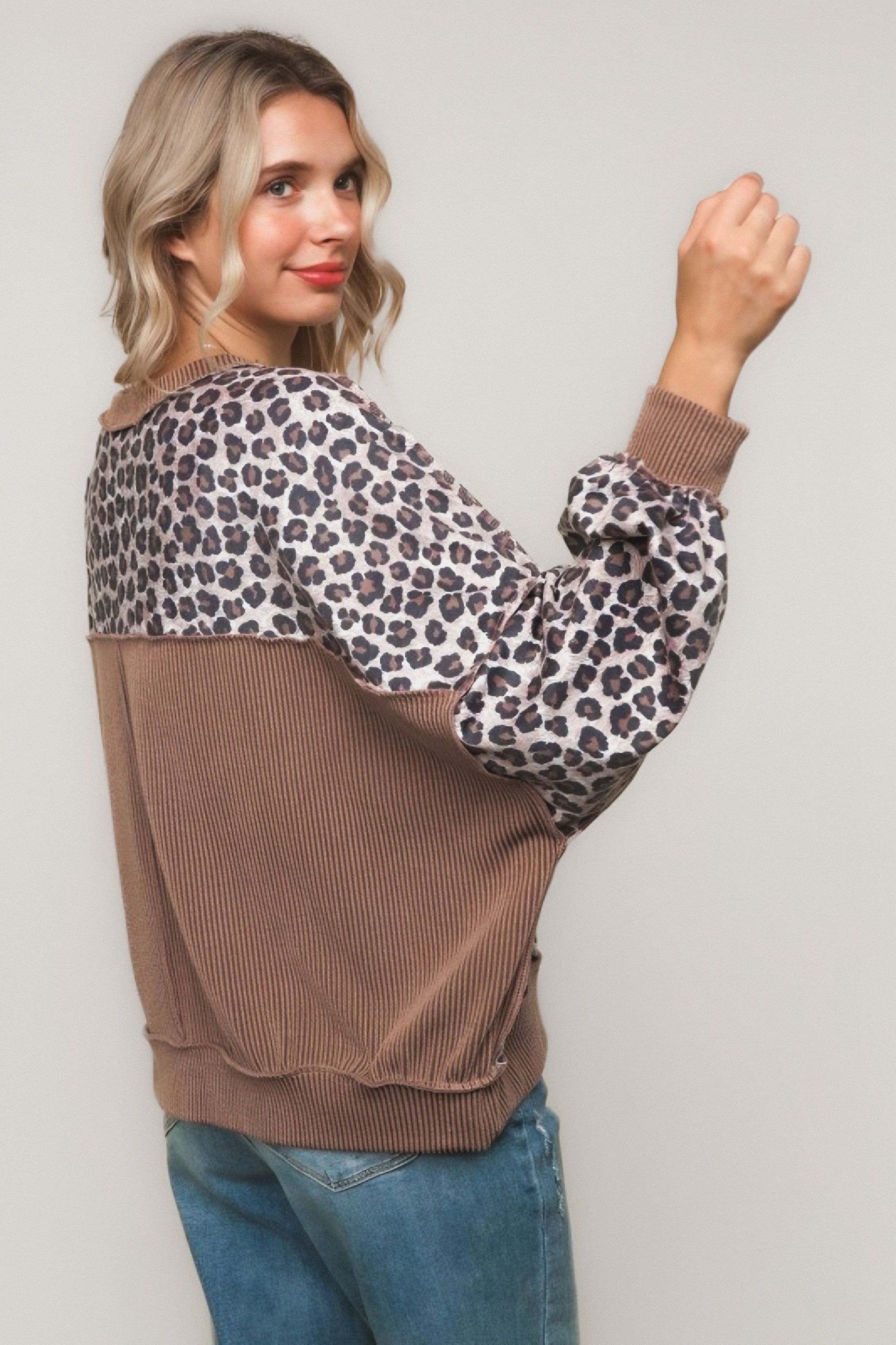 Woman with blonde hair wearing a brown pullover with leopard print sleeves and relaxed fit jeans against plain background