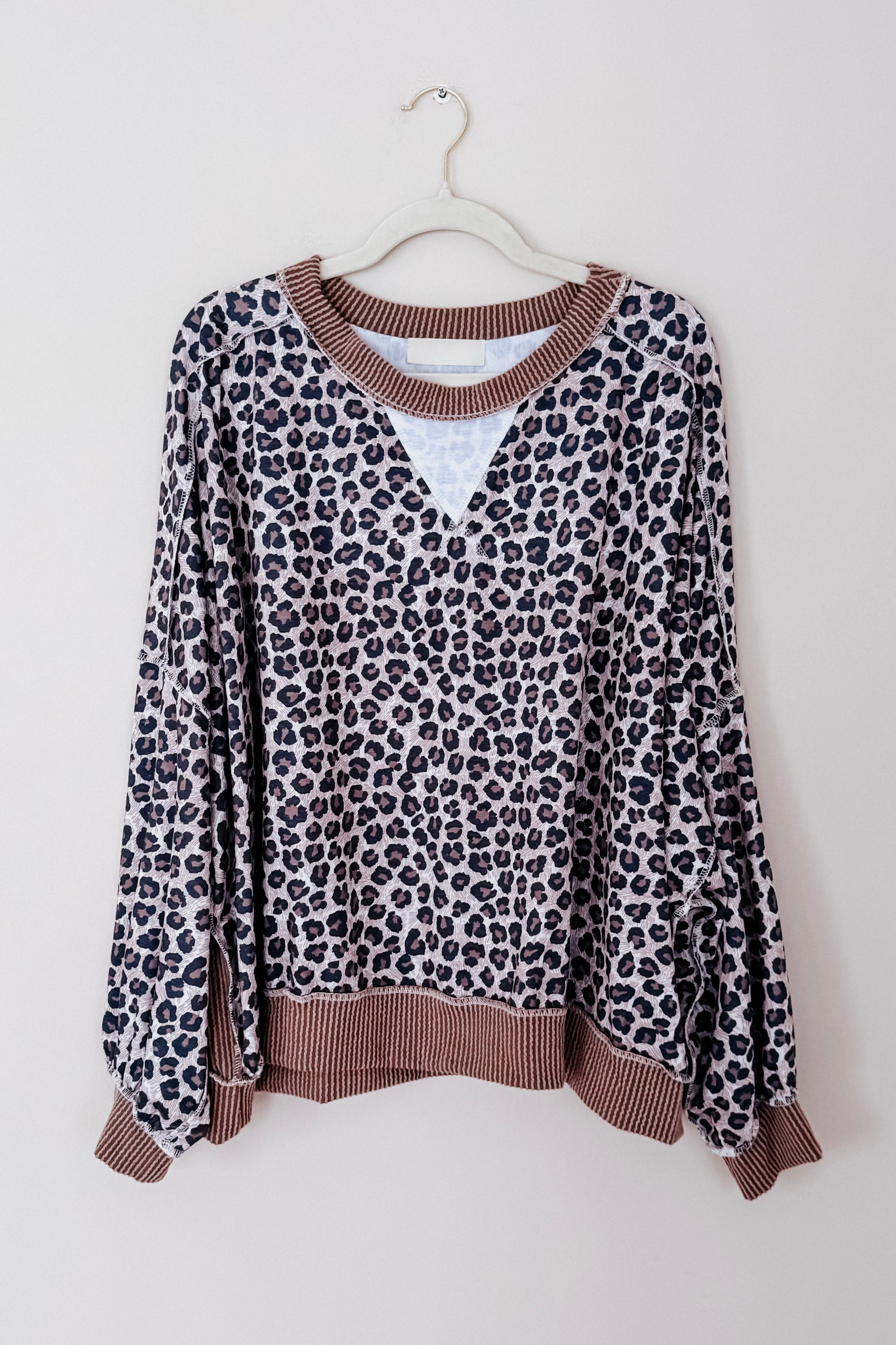 Leopard print pullover with brown ribbed cuffs, collar, and hem hanging on white hanger against plain wall