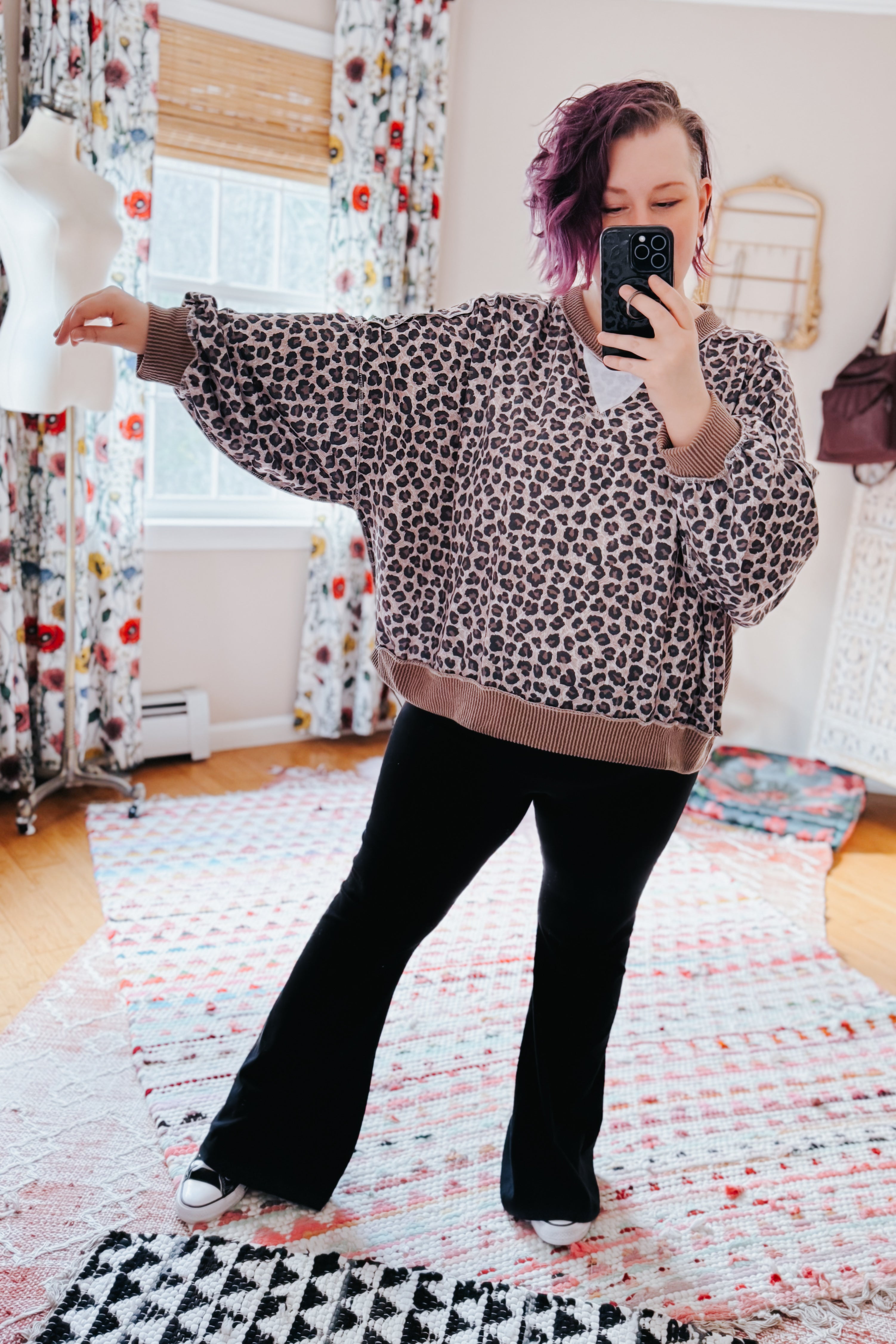 Woman with short purple hair wearing a brown leopard print cozy pullover and black pants taking a mirror selfie indoors