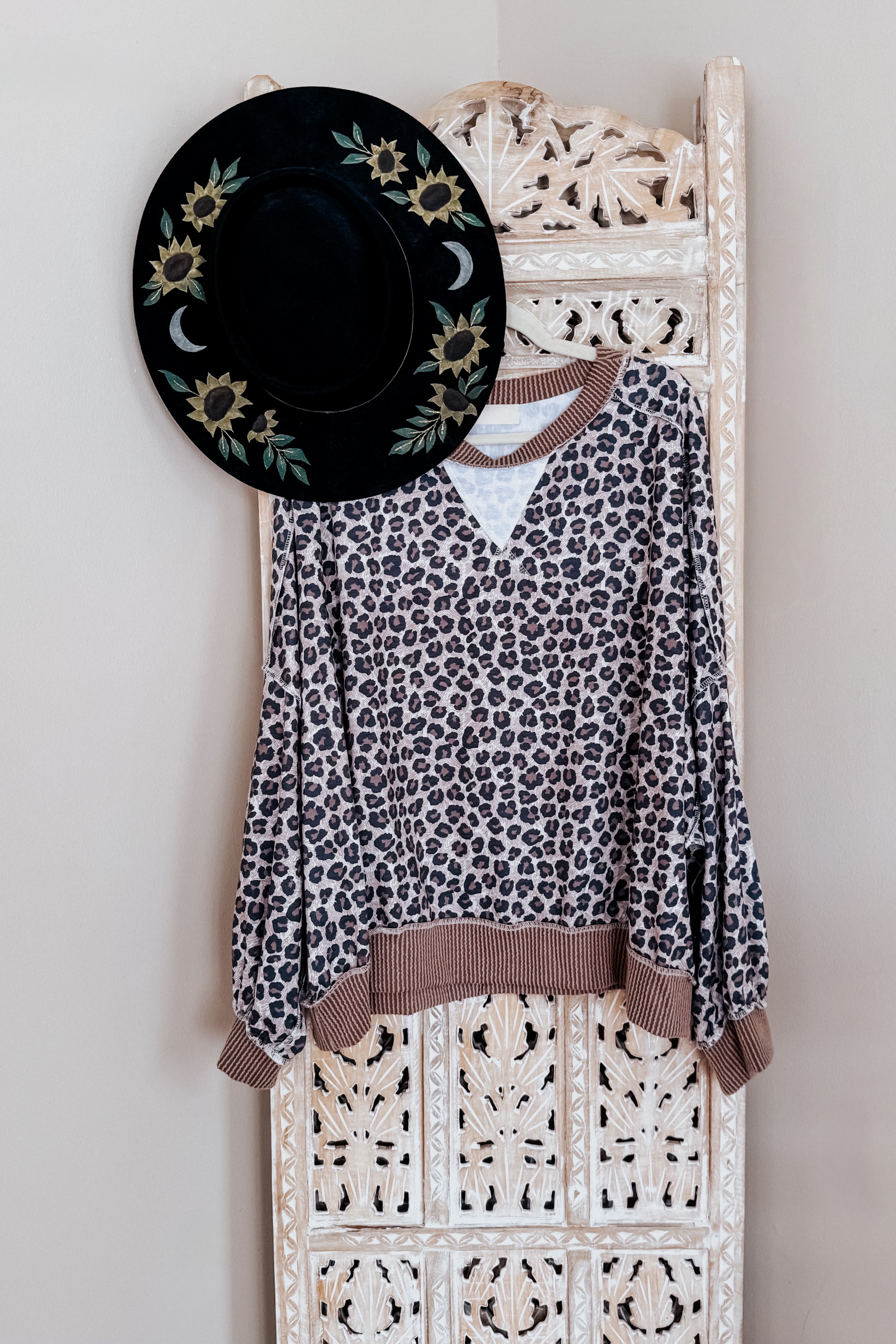 Brown leopard print pullover with ribbed cuffs and hem hanging on carved wooden panel beside black floral hat
