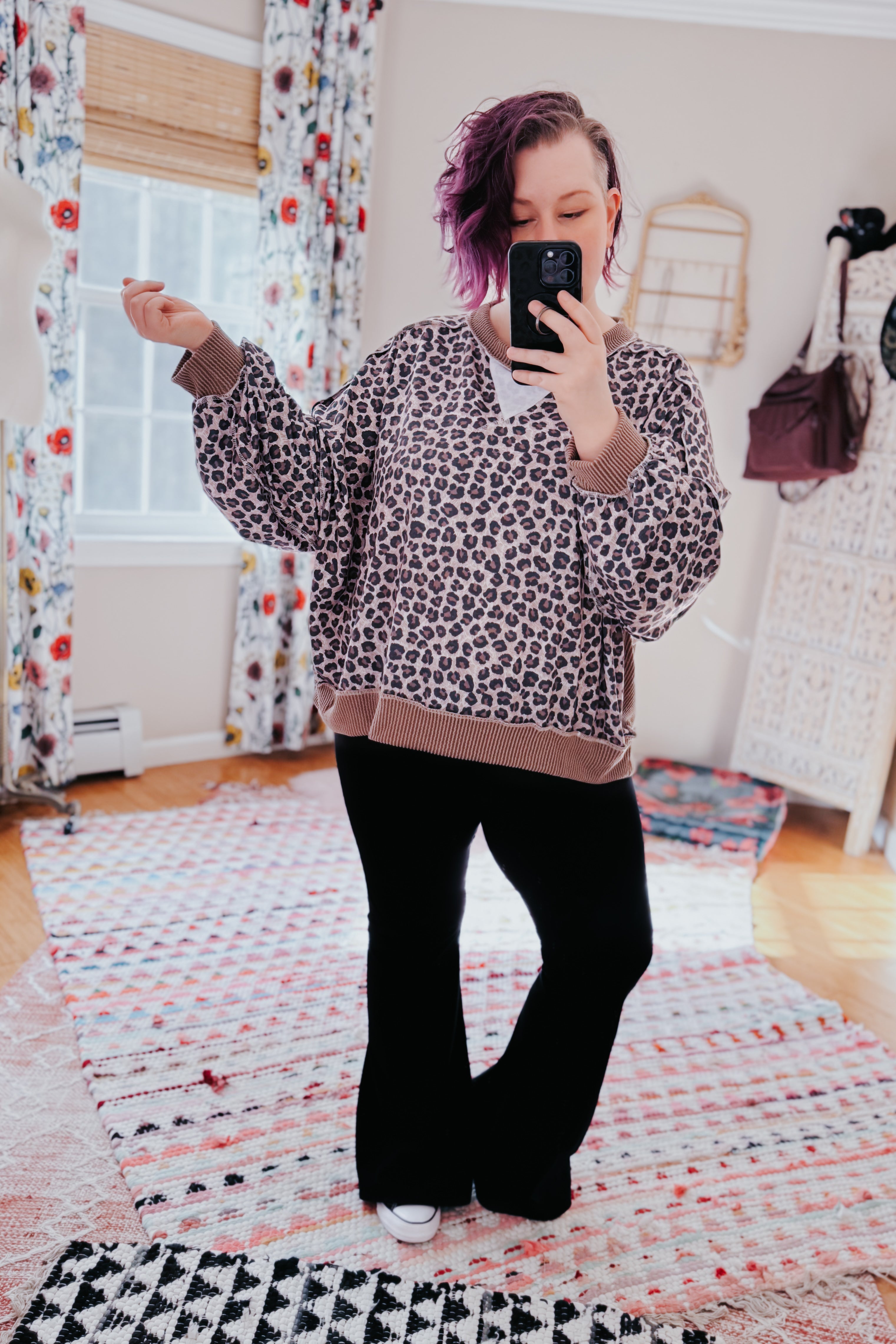 Woman with short dyed purple hair wearing a brown leopard print pullover and black pants taking a mirror selfie indoors