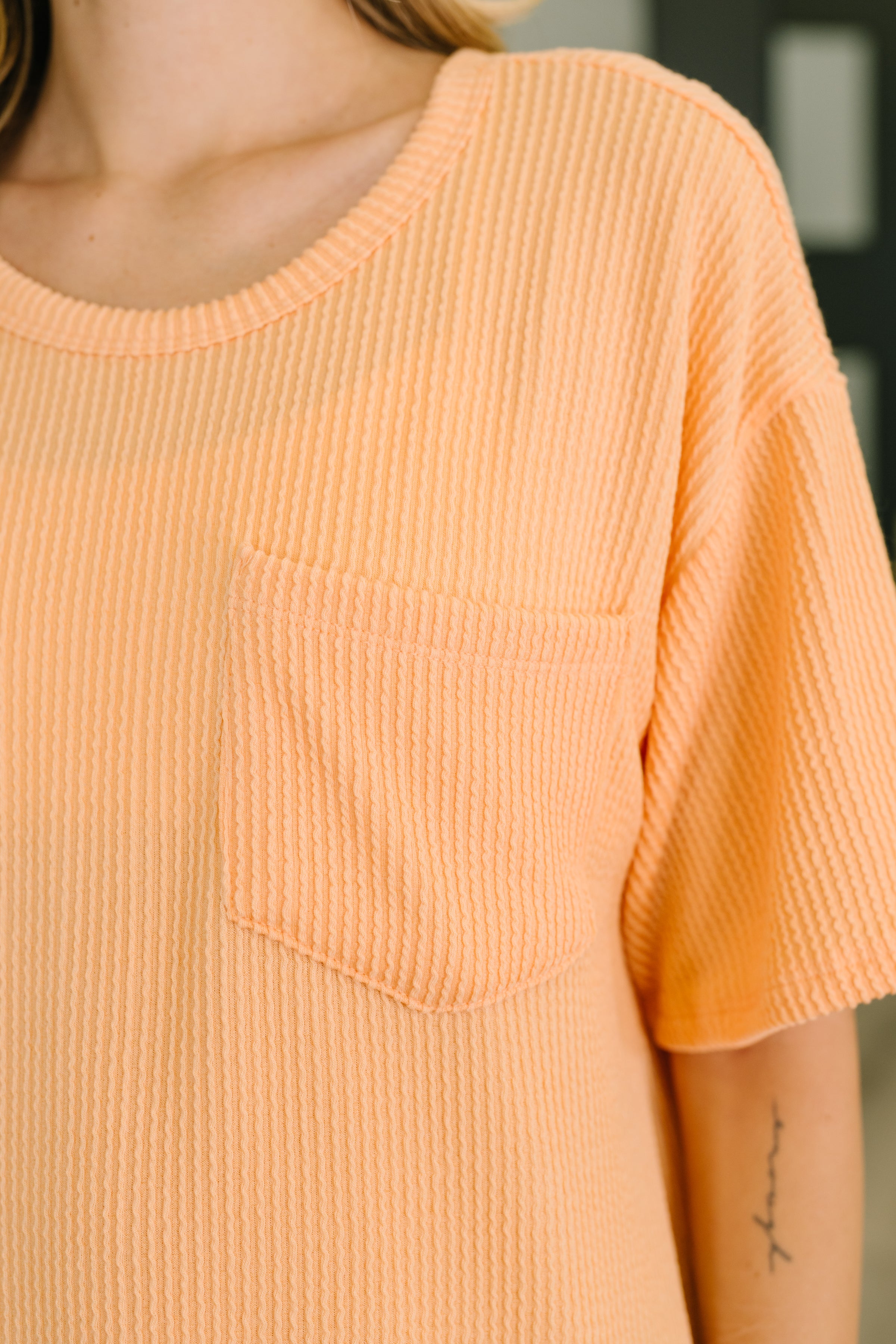 Close-up of relaxed fit orange textured tee with front pocket and scoop neckline on model indoors