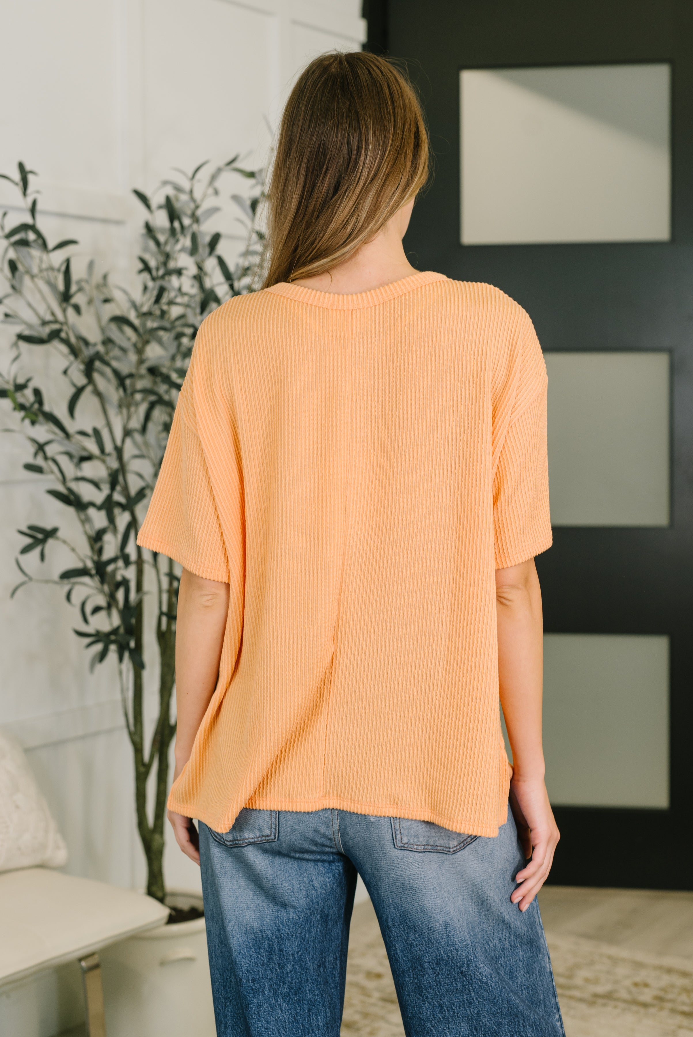 Back view of woman wearing loose apricot ribbed tee paired with relaxed fit blue jeans indoors