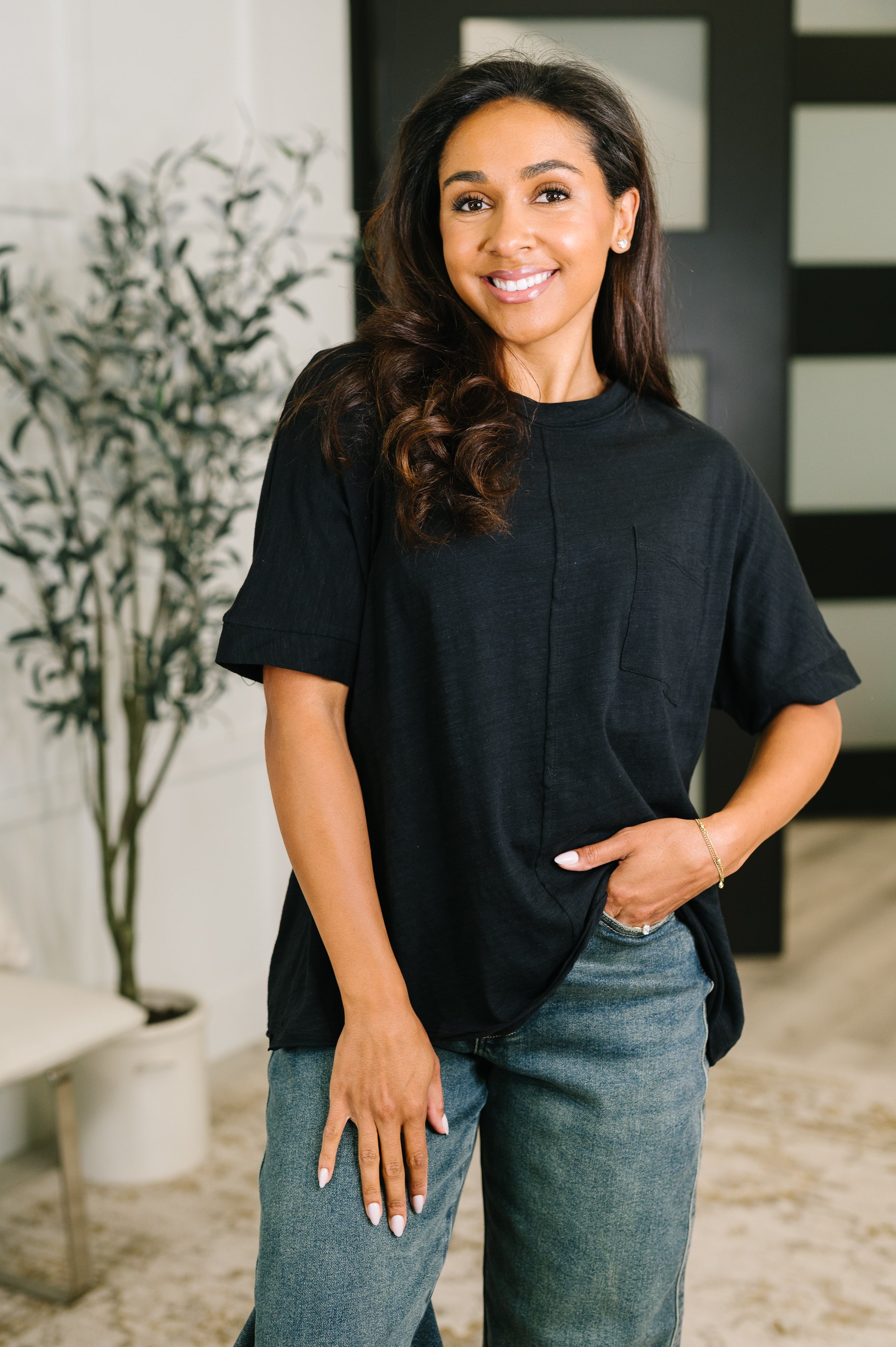 Smiling woman wearing a loose black boxy tee with rolled sleeves and faded blue jeans indoors with plant in background