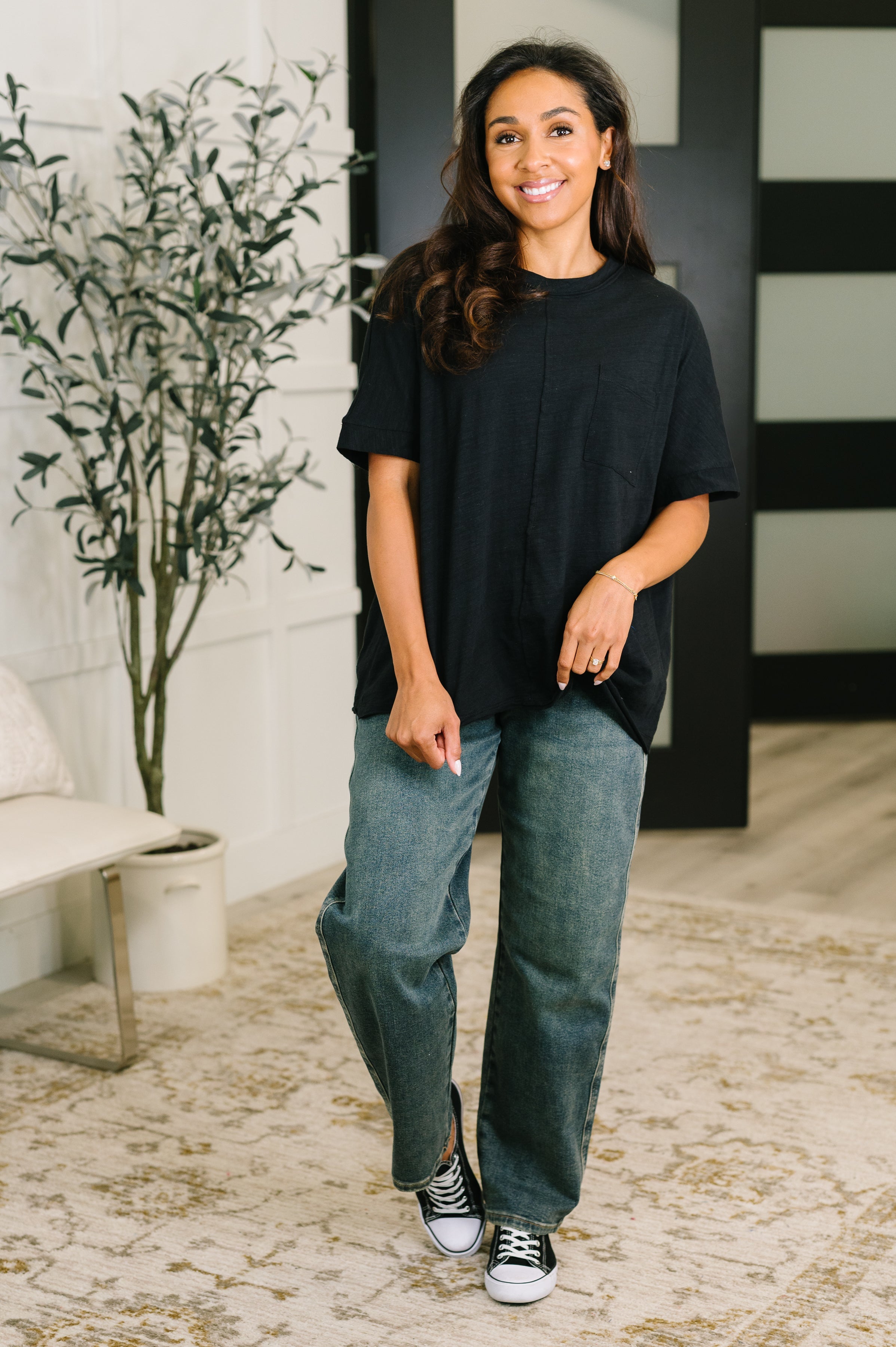 Smiling woman wearing a relaxed black tee with a subtle chest pocket paired with loose blue jeans and sneakers indoors