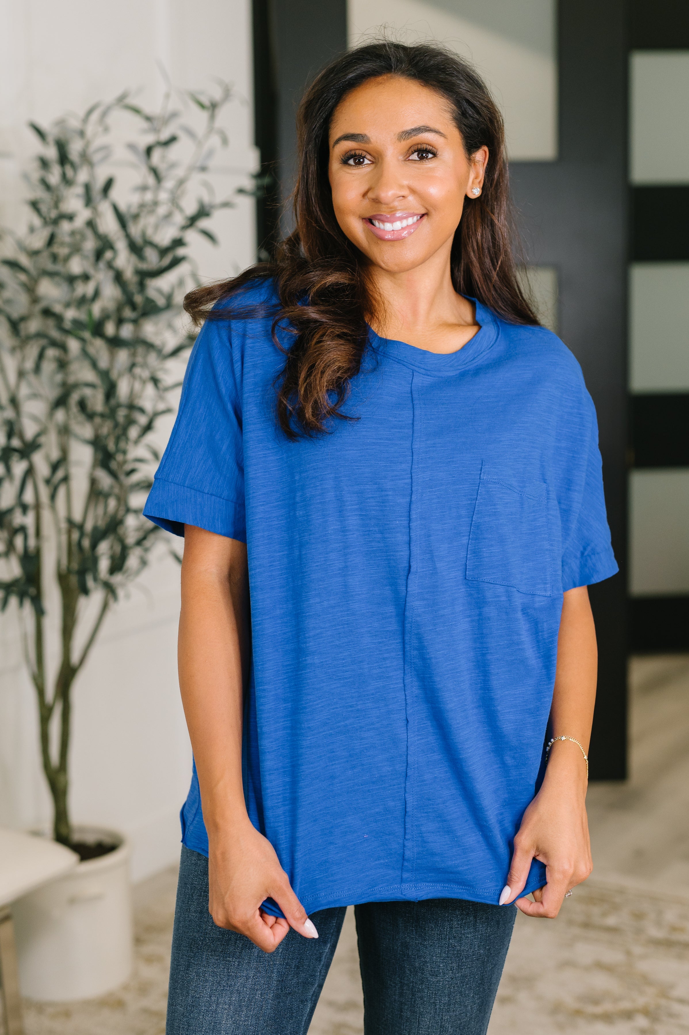 Smiling woman wearing a relaxed blue tee with a chest pocket and short sleeves standing indoors near plant