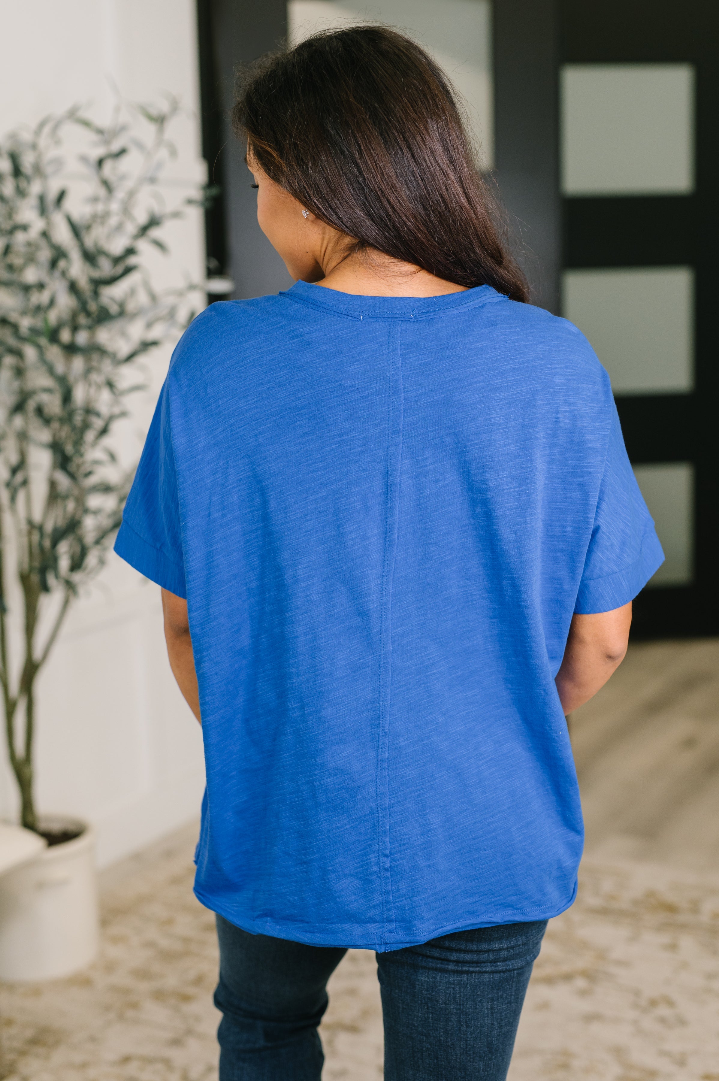 Back view of woman wearing relaxed fit blue tee with short sleeves and crew neck paired with dark jeans indoors