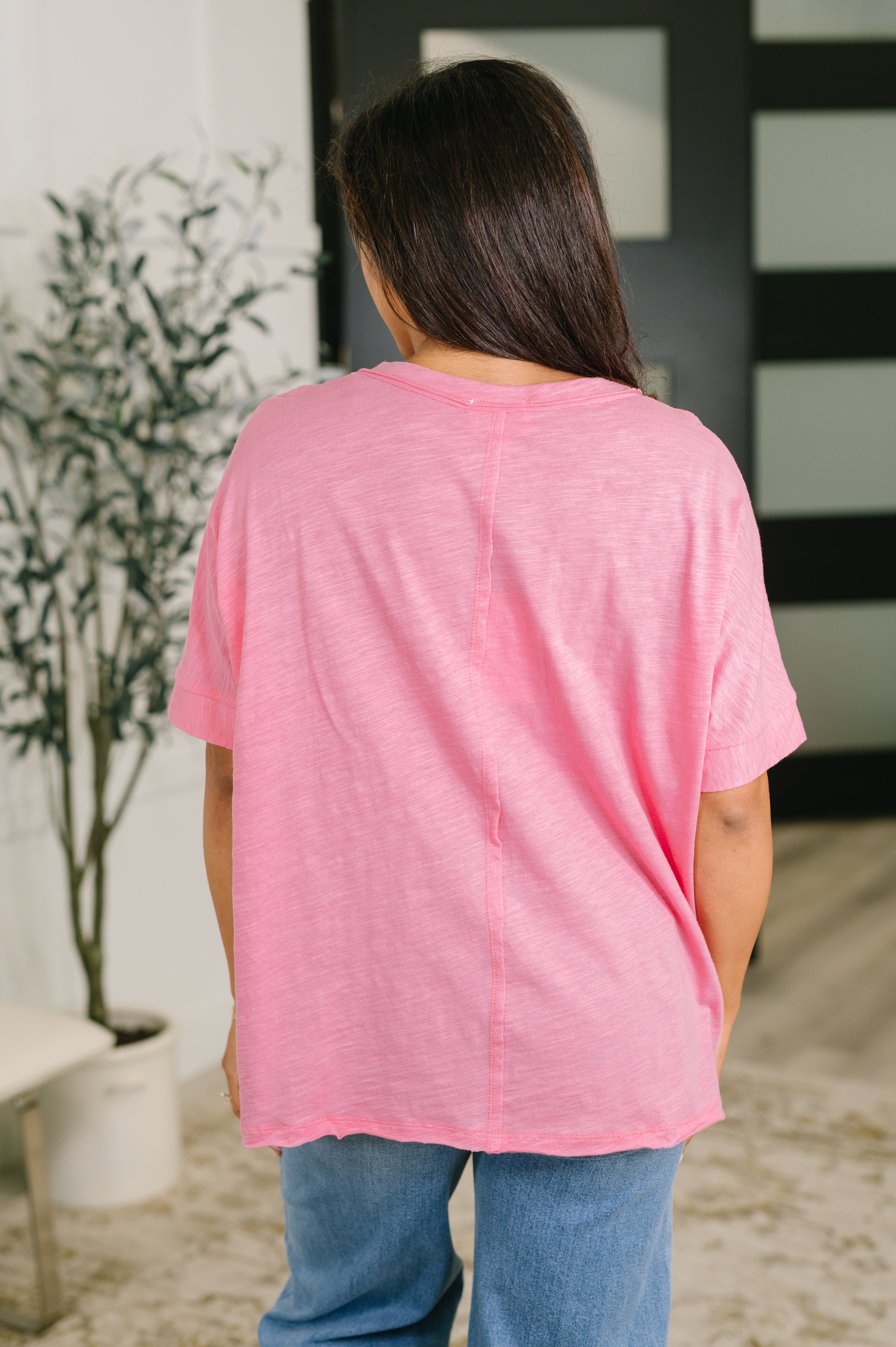 Back view of woman wearing loose pink heathered tee with rolled sleeves paired with blue jeans indoors
