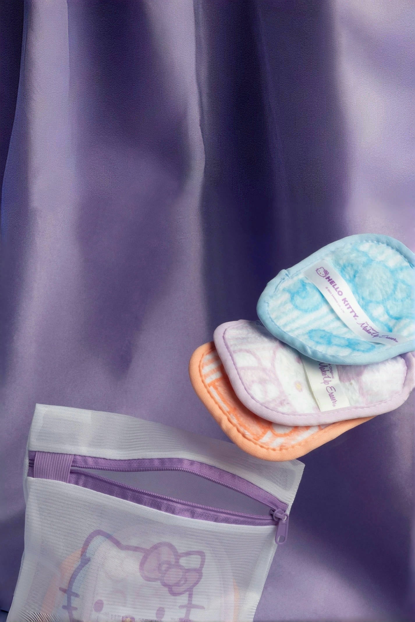 Soft reusable makeup remover pads in orange, blue, and purple with Hello Kitty pouch against purple fabric background