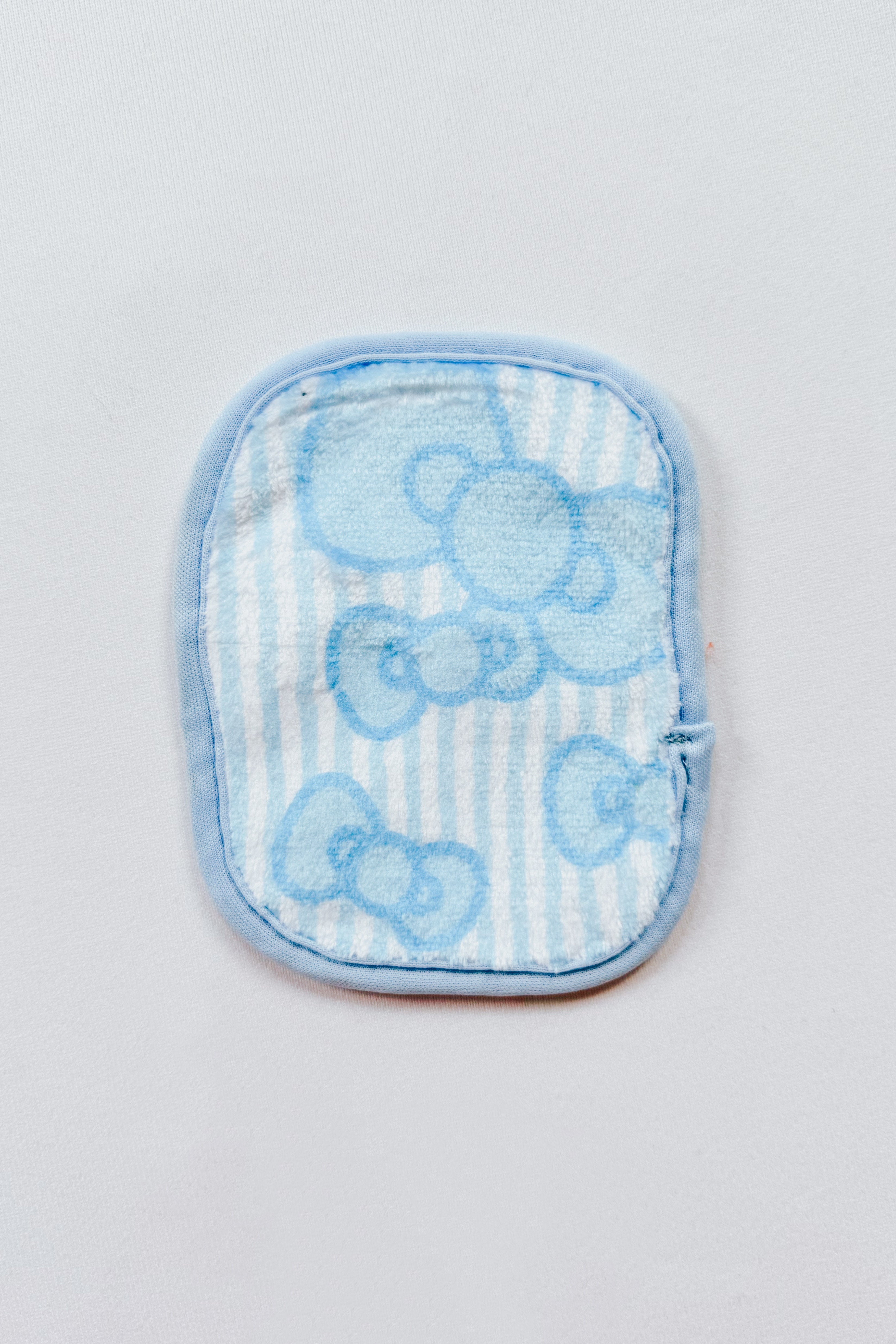 Small rectangular makeup remover pad with light blue trim and subtle Hello Kitty bow pattern on striped fabric