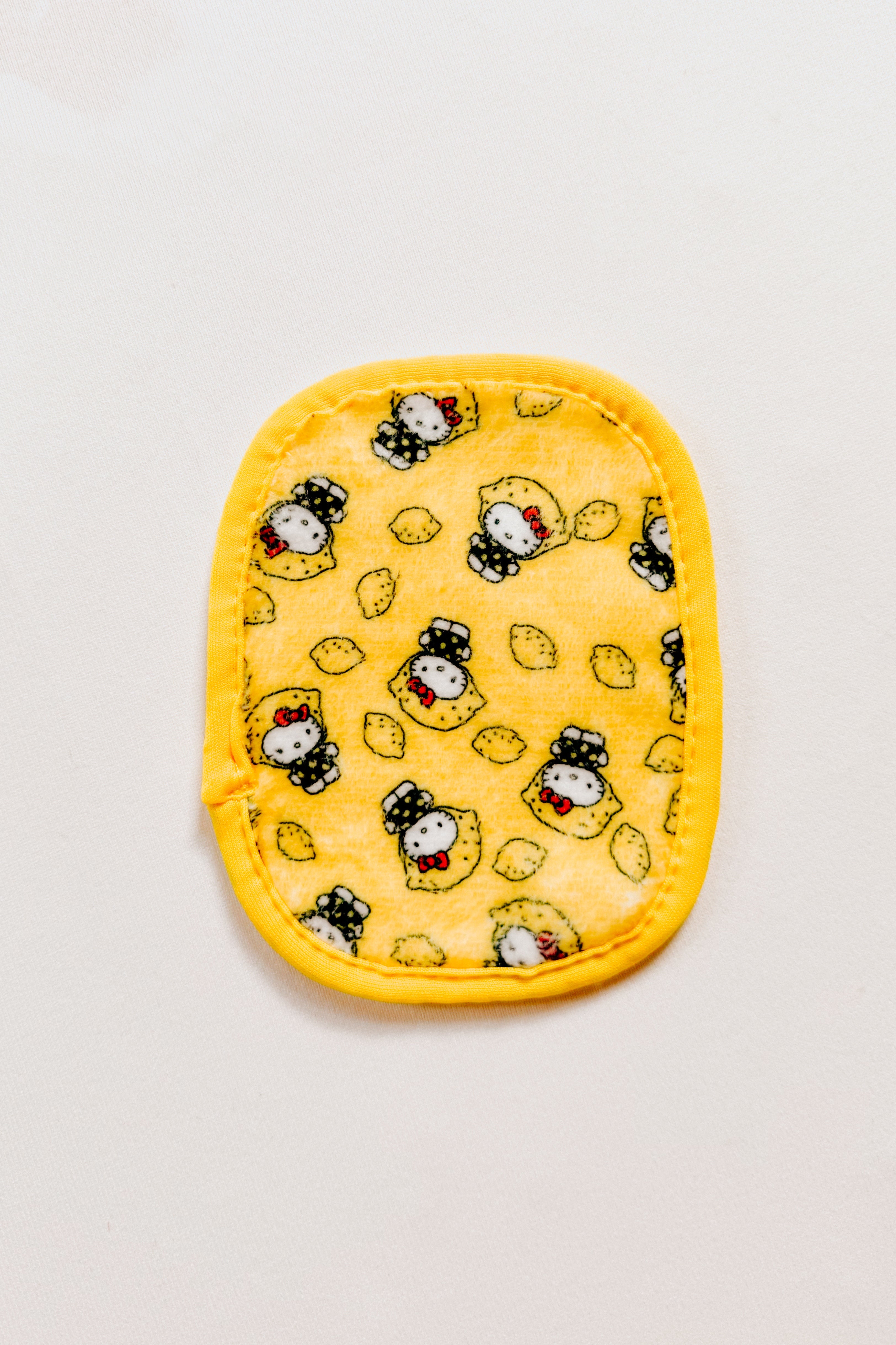Small yellow reusable makeup eraser pad featuring playful Hello Kitty faces in black and white with lemon motifs