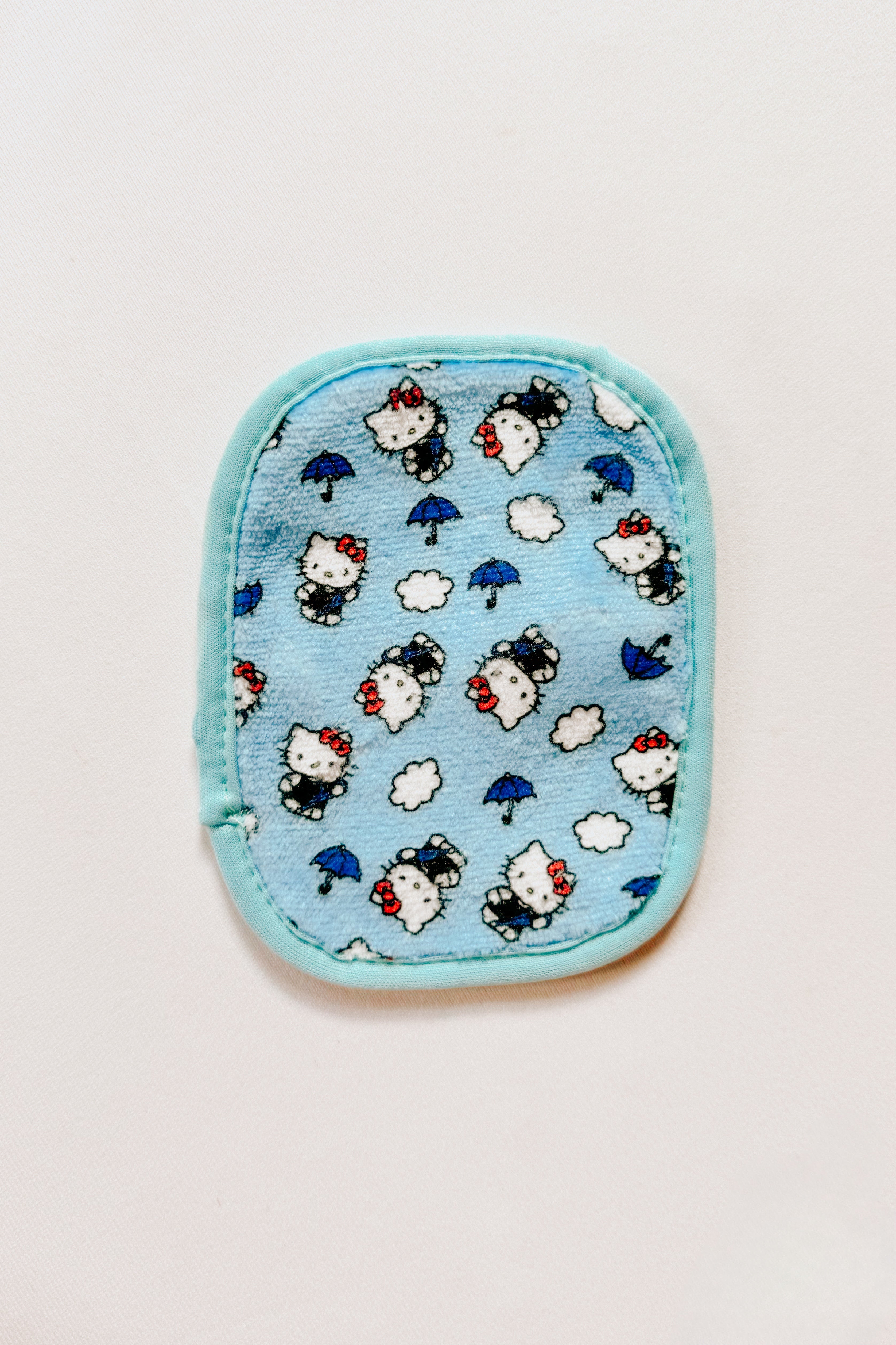 Small blue makeup eraser pad featuring playful Hello Kitty characters, umbrellas, and clouds on soft fabric background