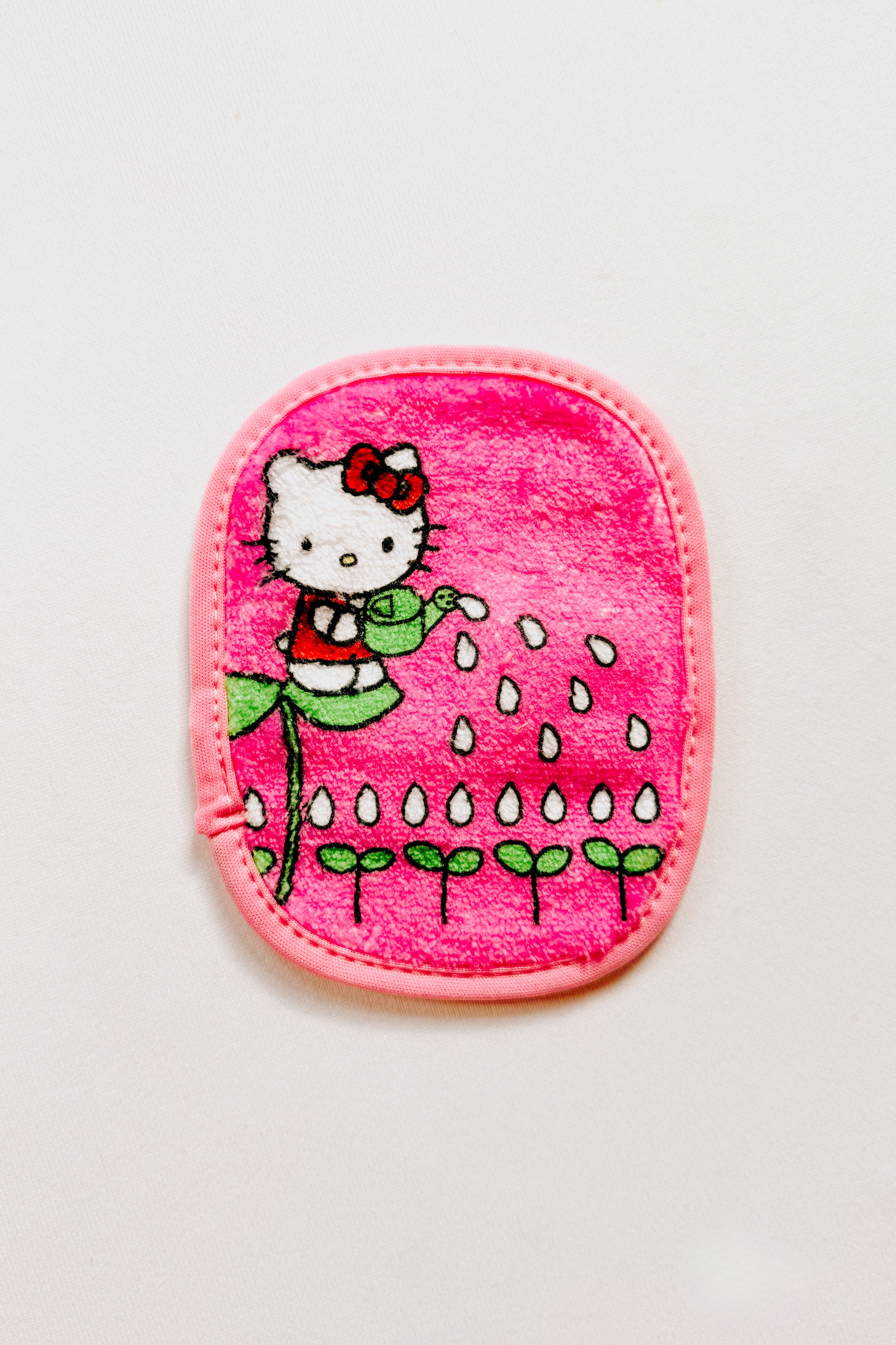 Small pink makeup remover pad featuring Hello Kitty watering plants with water droplets on bright background