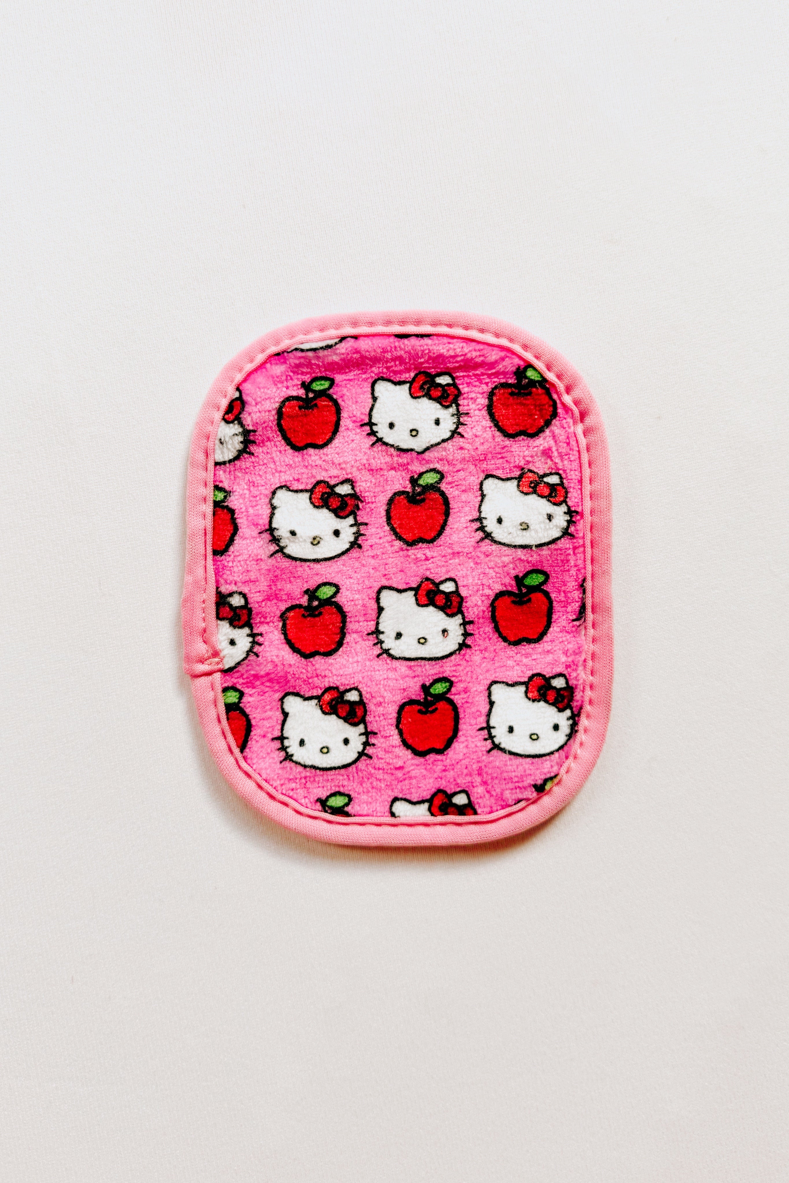 Small pink makeup eraser cloth with red apples and cartoon cat faces pattern featuring bows on white background