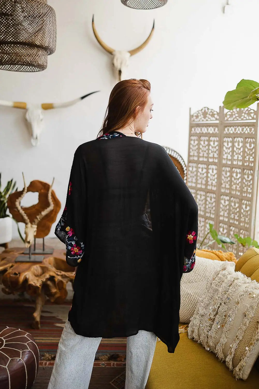 Woman wearing a black embroidered duster with floral details on sleeves in a cozy eclectic living room setting