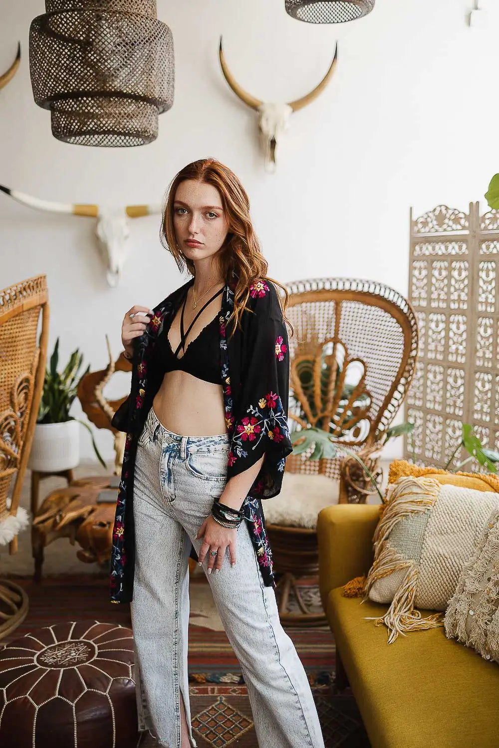 Woman wearing black embroidered duster over bralette paired with light wash jeans in eclectic living room setting