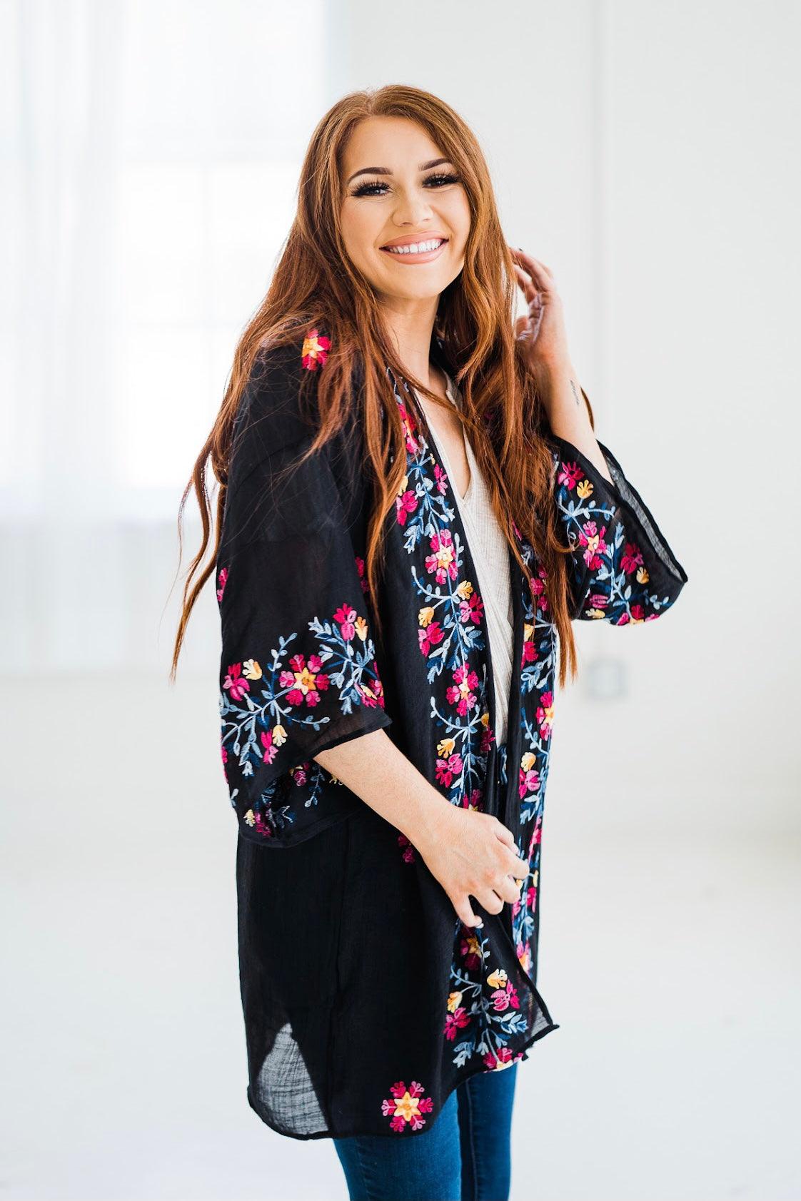 Smiling woman wearing a black embroidered duster with colorful floral patterns in a bright indoor setting