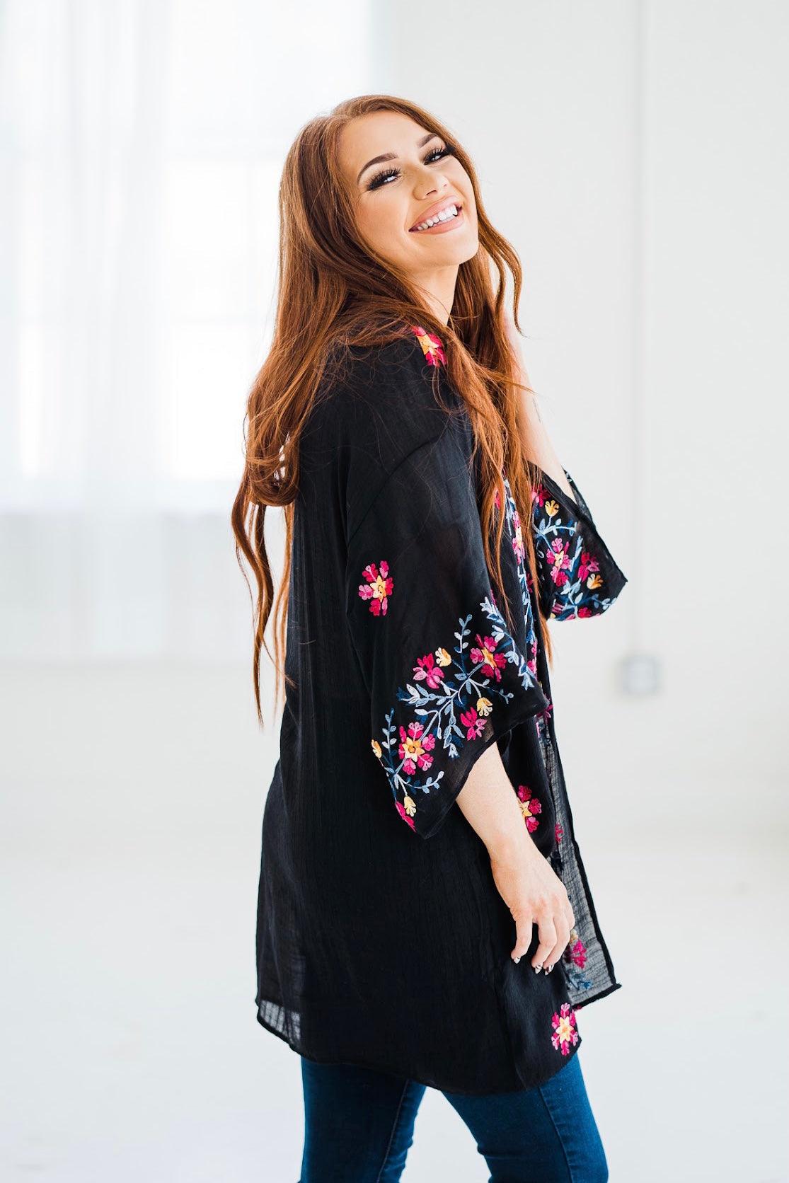 Smiling woman wearing black embroidered duster with colorful floral designs on sleeves and hem in bright indoor space