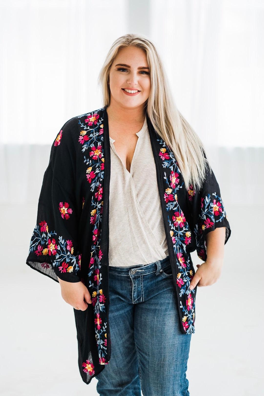 Smiling woman wearing a black embroidered duster with colorful floral details paired with casual jeans and blouse