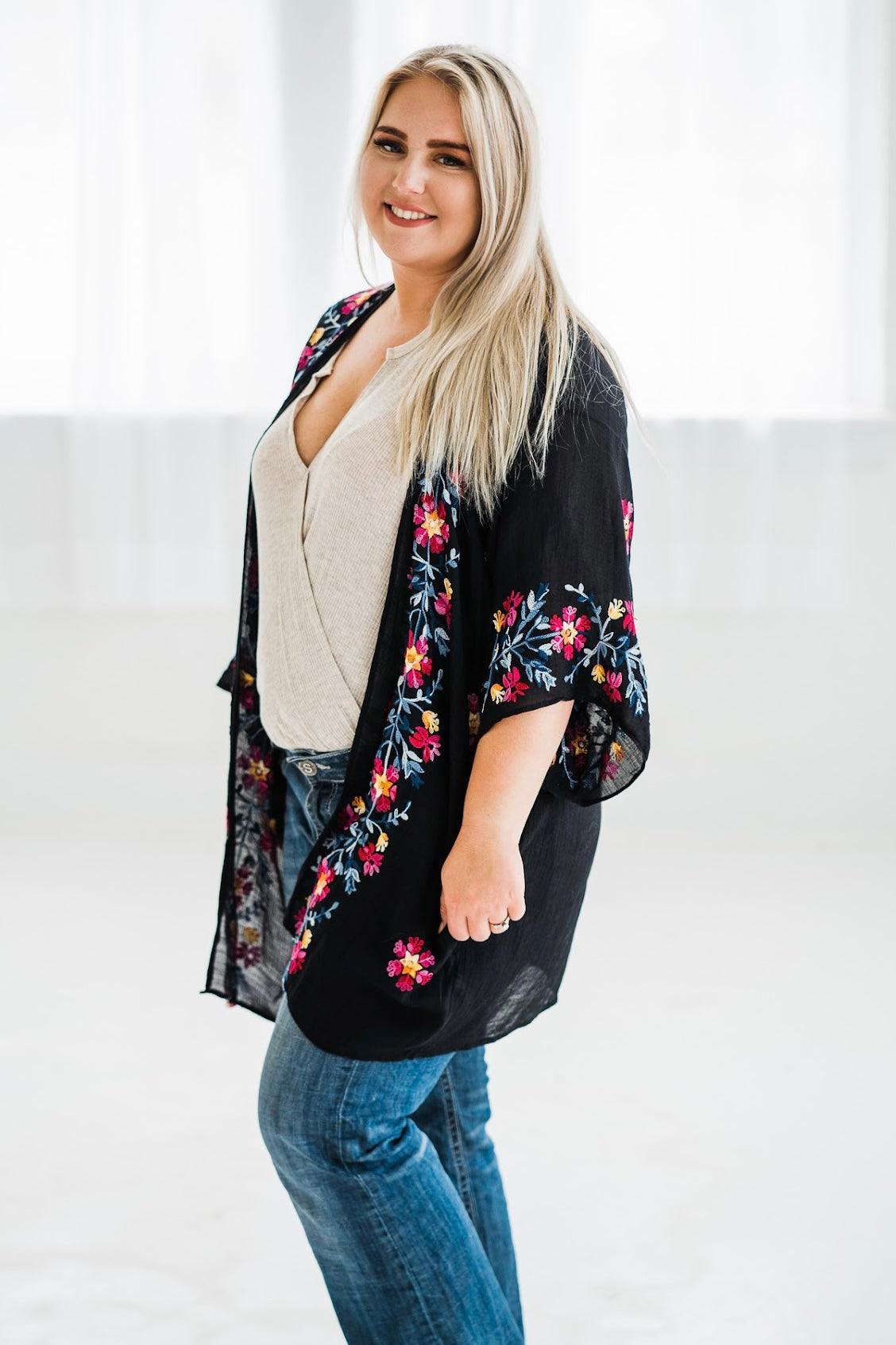 Smiling woman wearing black embroidered duster with floral details over beige top and blue jeans in bright studio