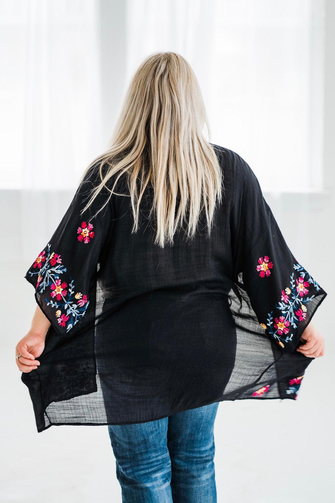 Back view of a size-inclusive black embroidered duster with colorful floral details worn with blue jeans indoors