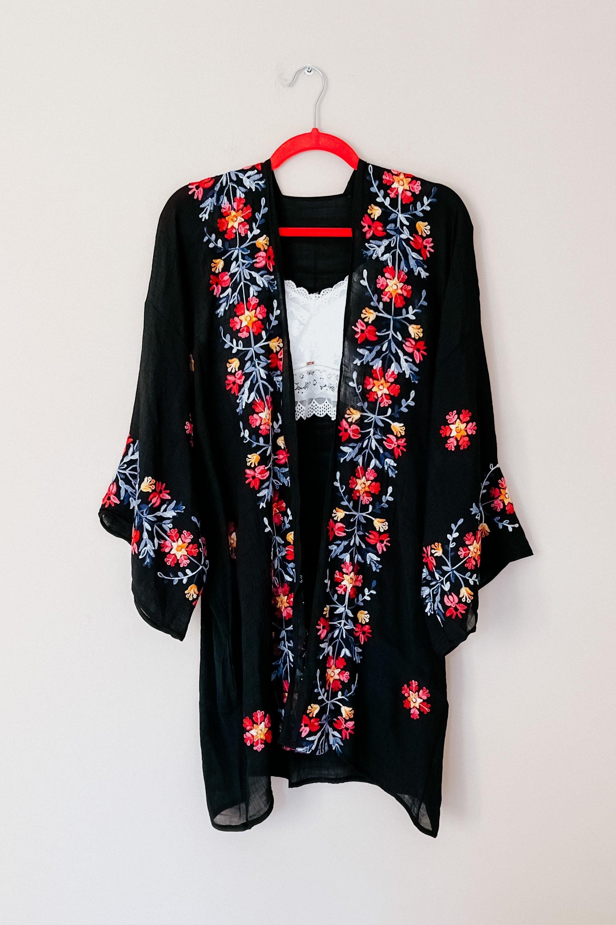 Black duster with colorful floral embroidery on sleeves and front, hanging on a red hanger against a white wall