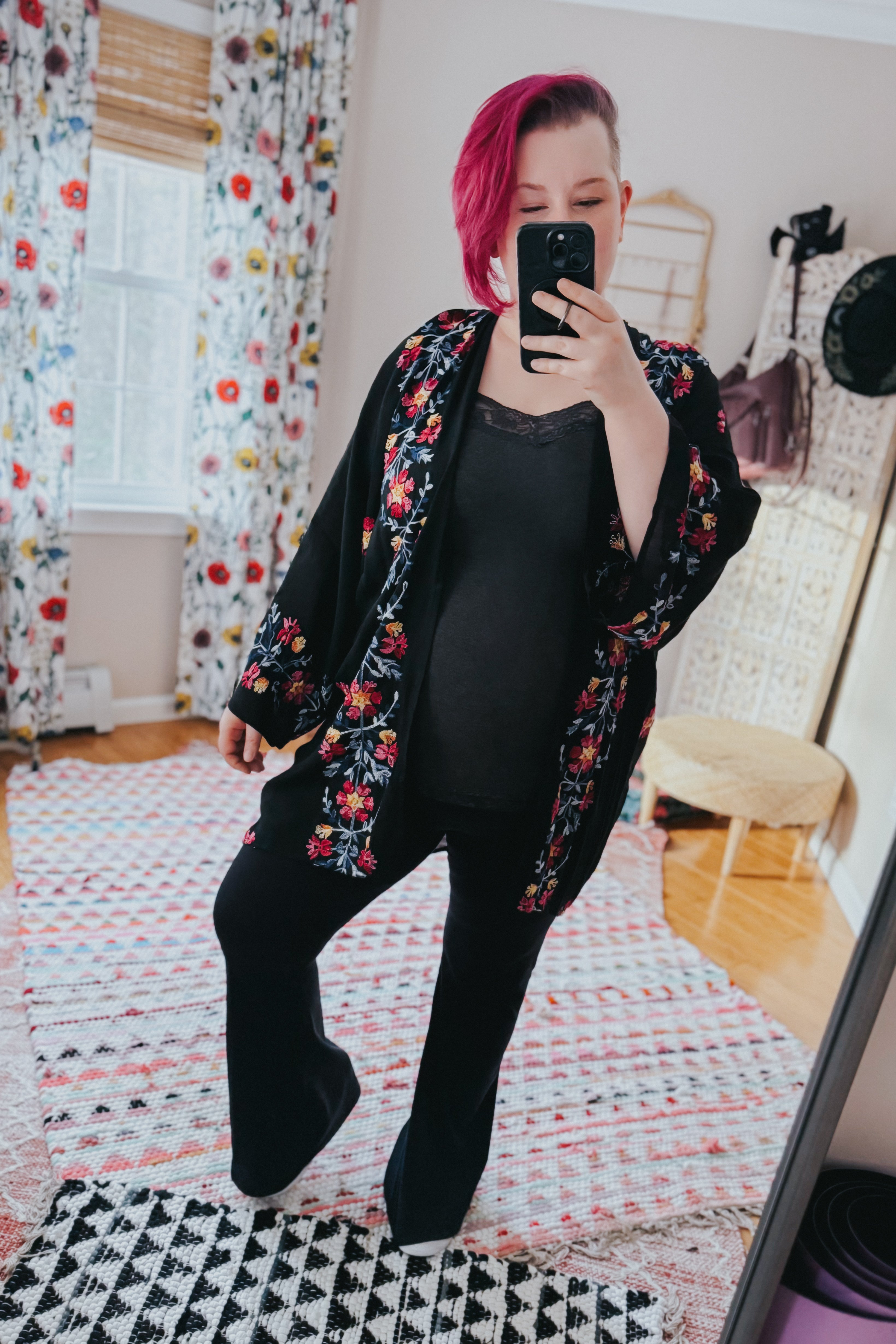 Woman with magenta hair wearing black embroidered duster and black outfit taking mirror selfie in bright room with floral curtains