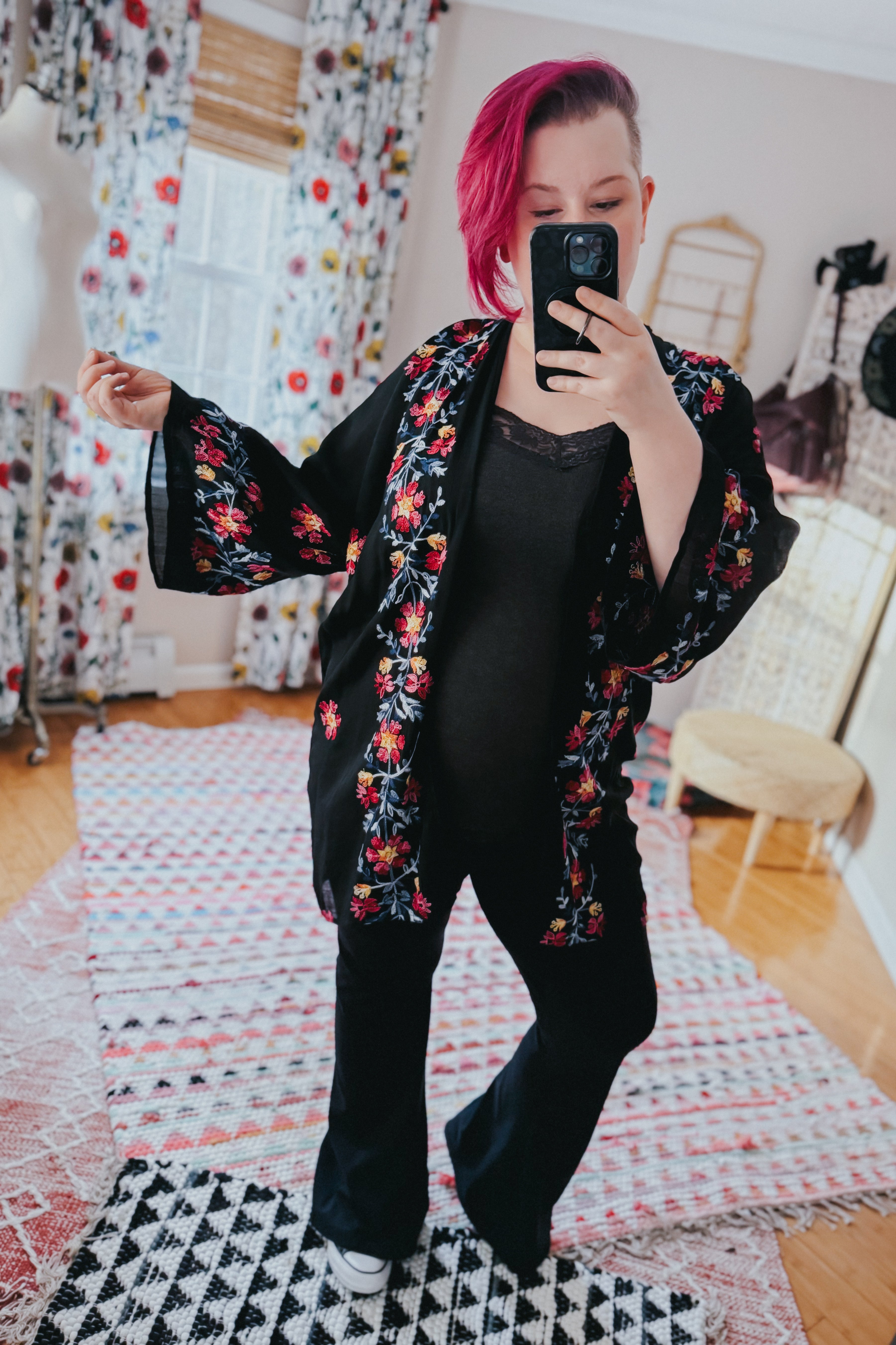 Woman with pink hair wearing black embroidered duster with floral pattern over black outfit taking selfie indoors