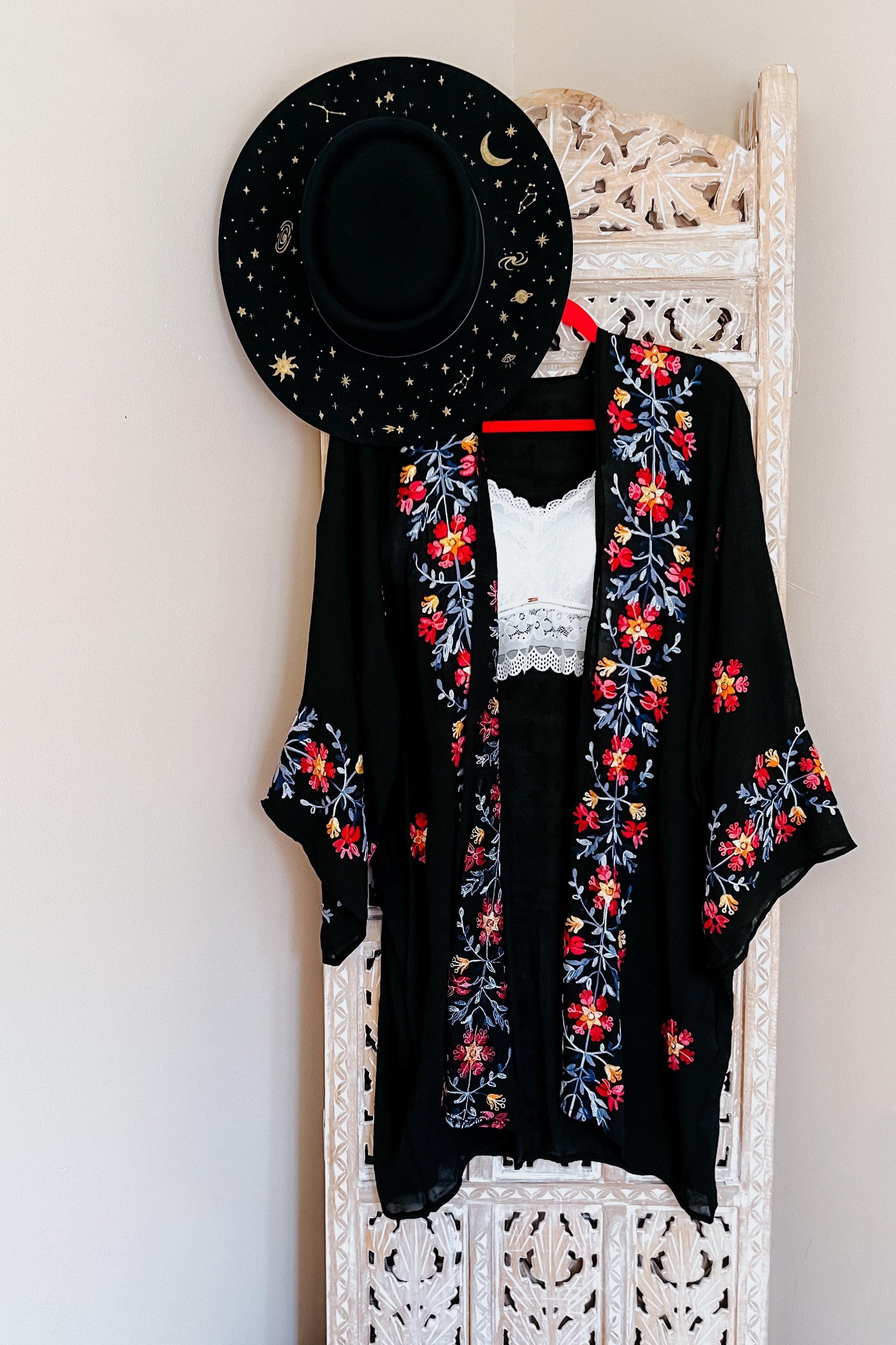 Black embroidered duster with vibrant floral patterns hanging on carved wooden panel with black starry wide-brim hat