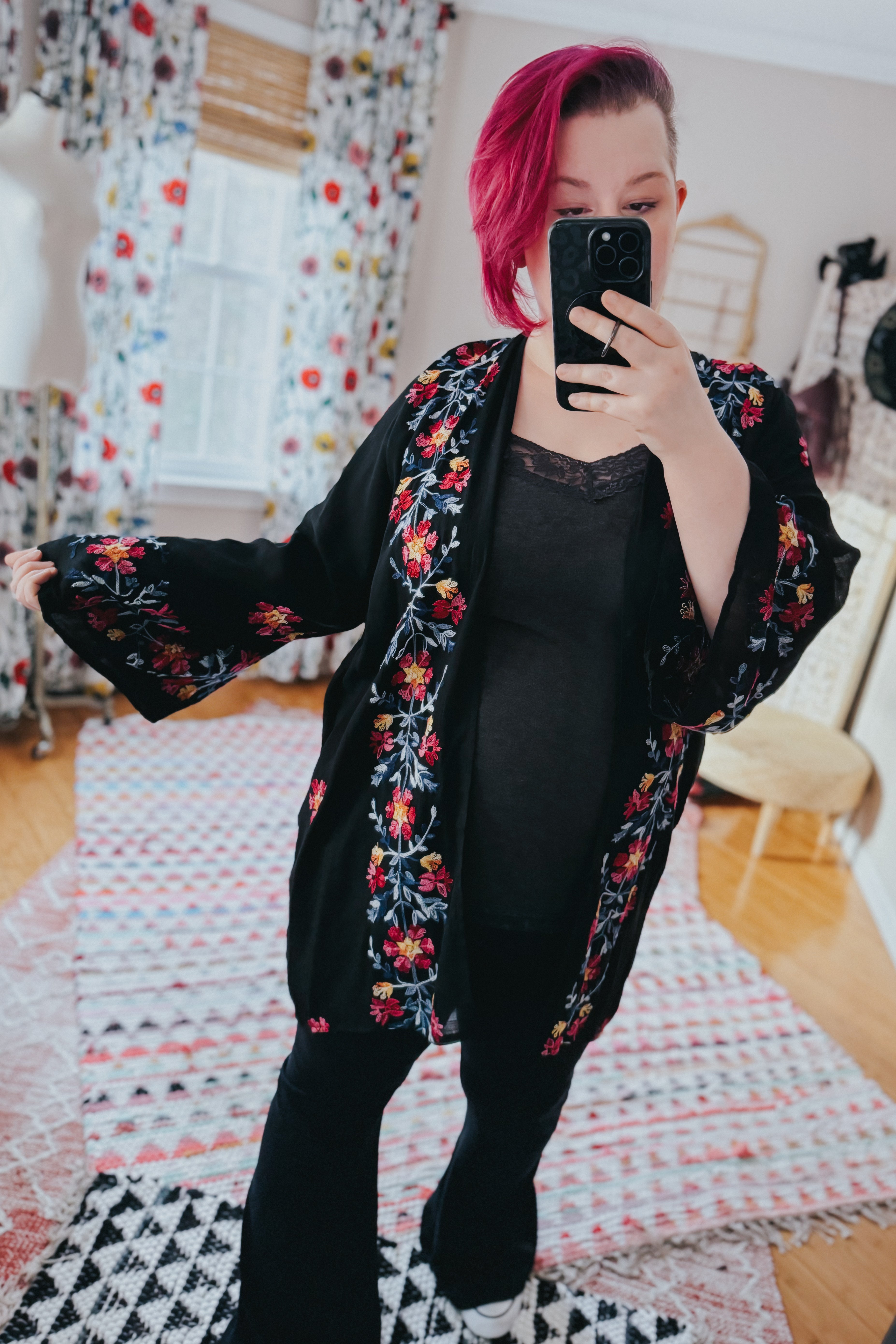 Woman wearing black embroidered duster with floral details and wide sleeves taking a mirror selfie indoors
