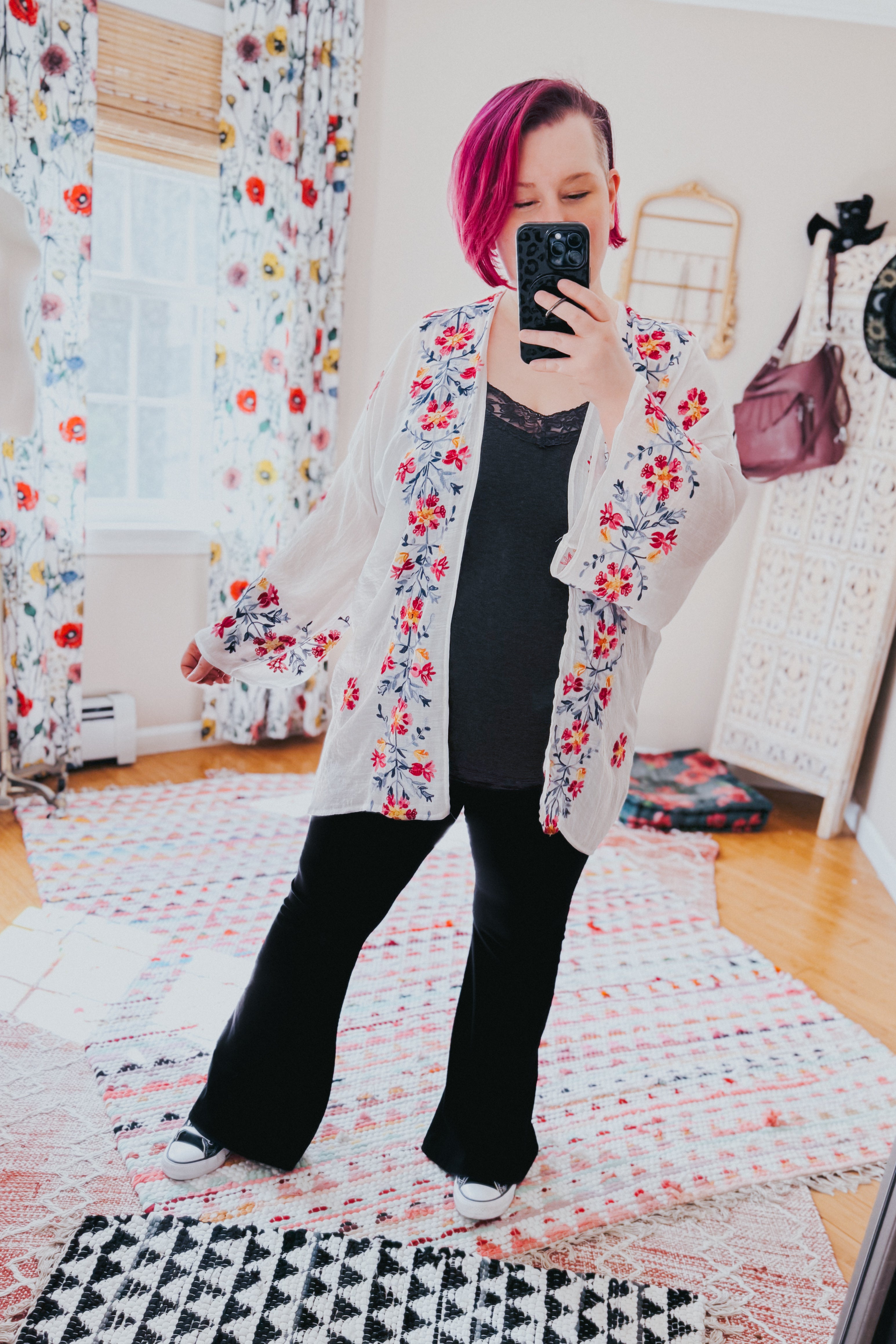 Woman wearing an ivory embroidered duster with floral details over black top and pants in a bright eclectic room