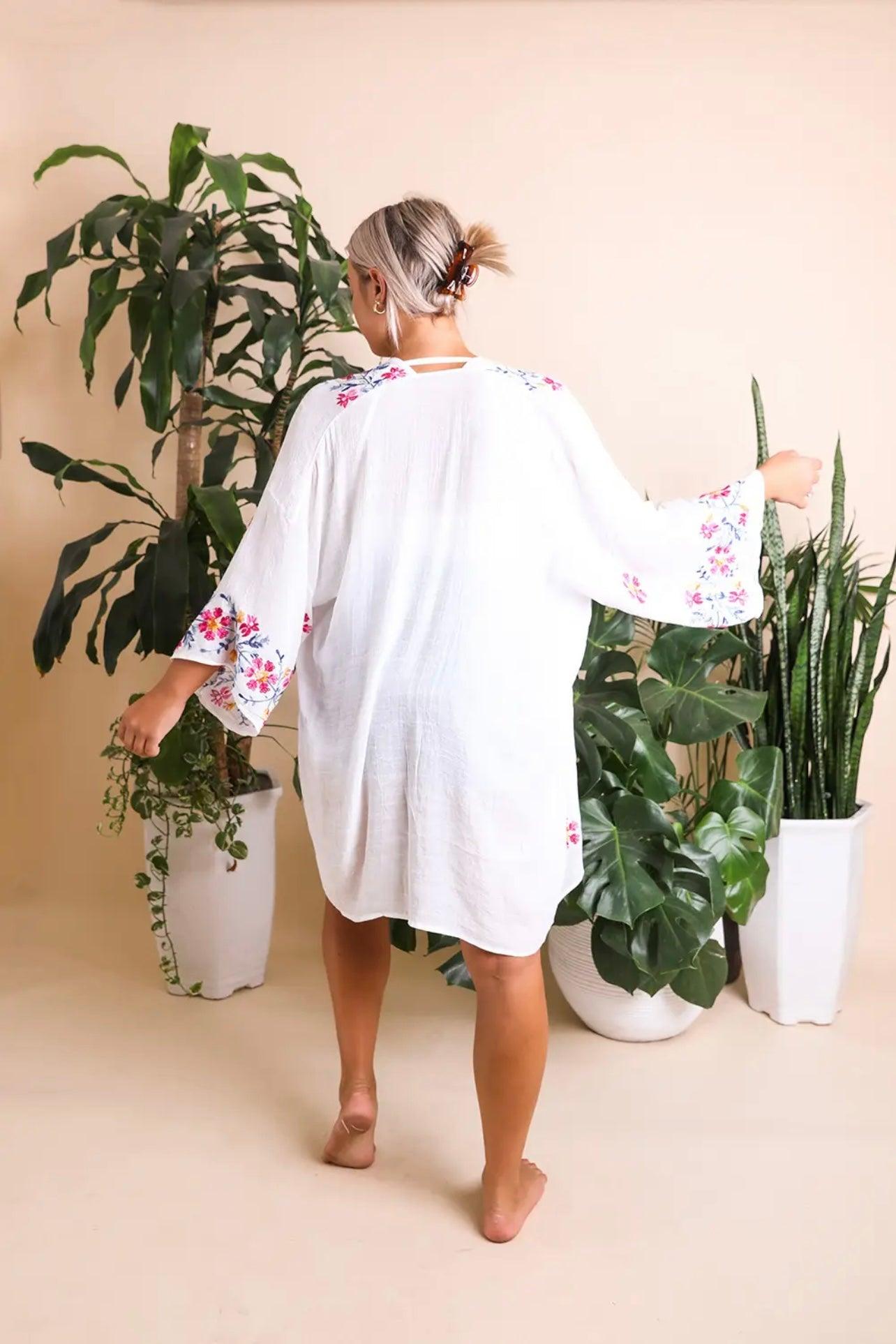 Woman wearing an ivory embroidered duster with floral accents on sleeves and back, standing barefoot among green plants