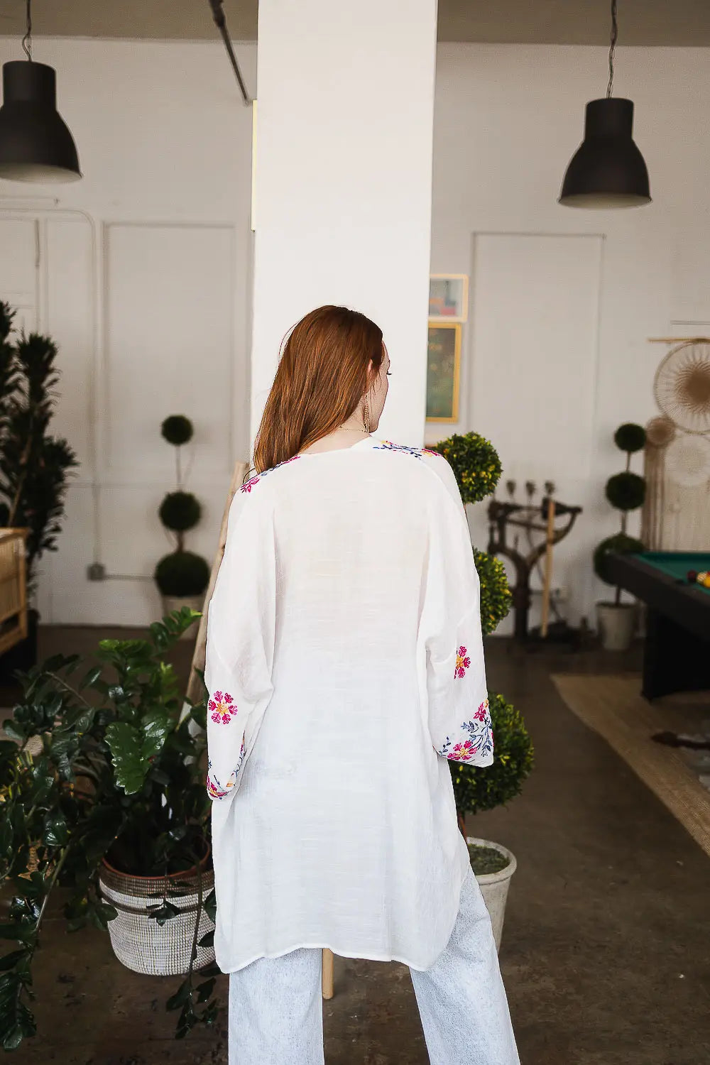 Woman with red hair wearing an ivory embroidered duster with floral accents, standing in a bright, cozy room
