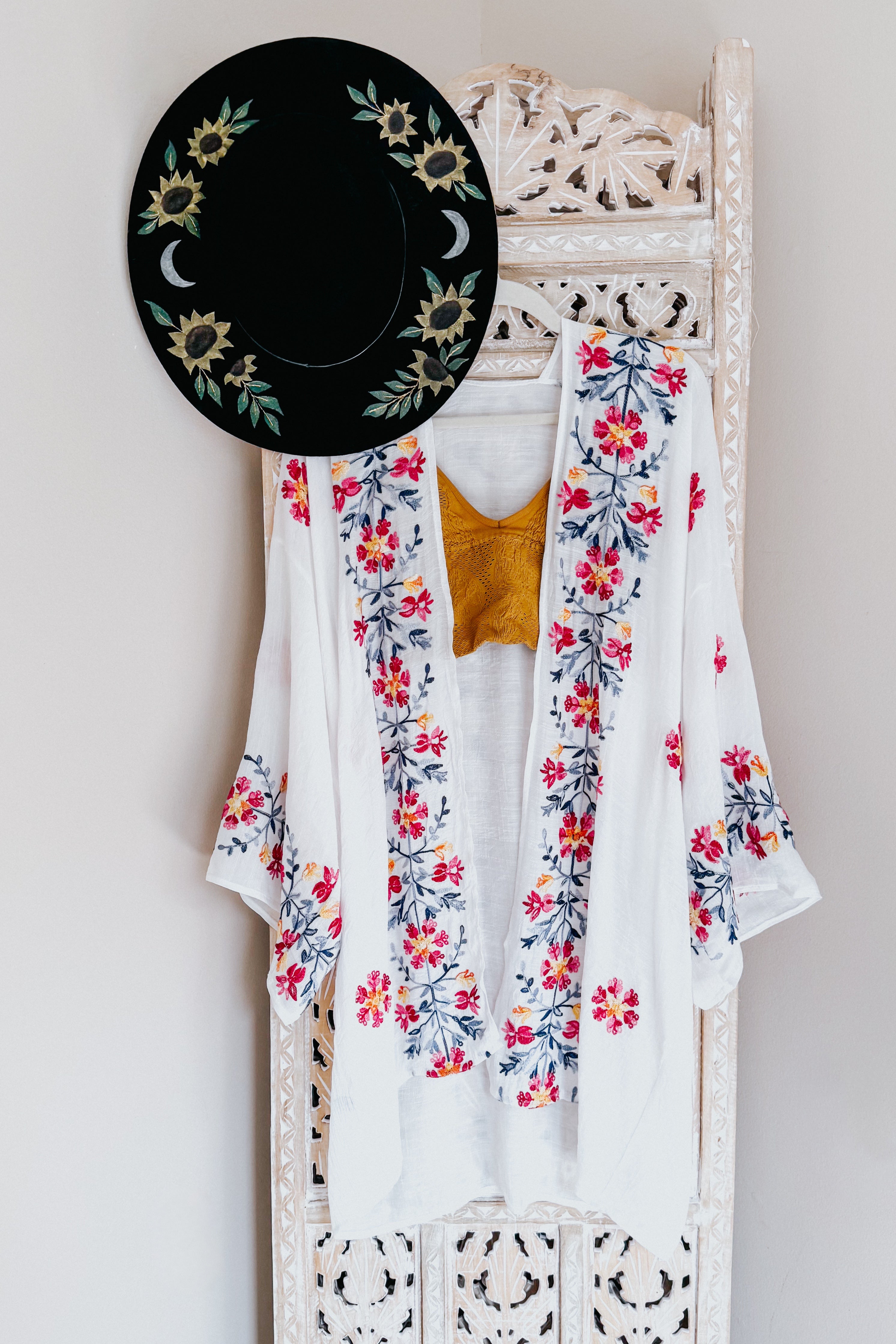 Ivory embroidered duster with floral designs layered over mustard top, paired with black hat featuring sunflower and moon patterns