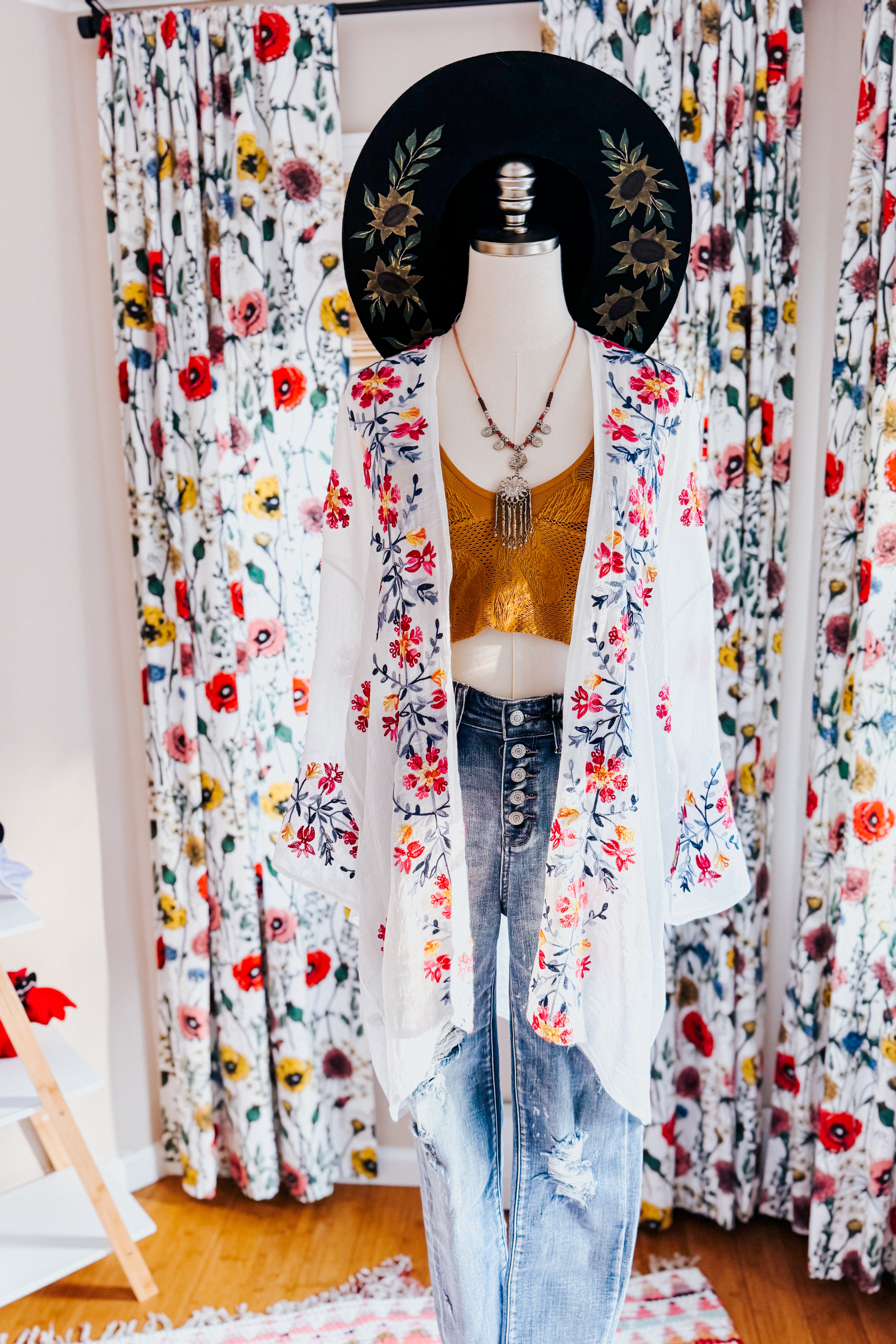 Mannequin dressed in an embroidered ivory duster over a mustard top paired with high-waisted distressed jeans and a black hat with floral details