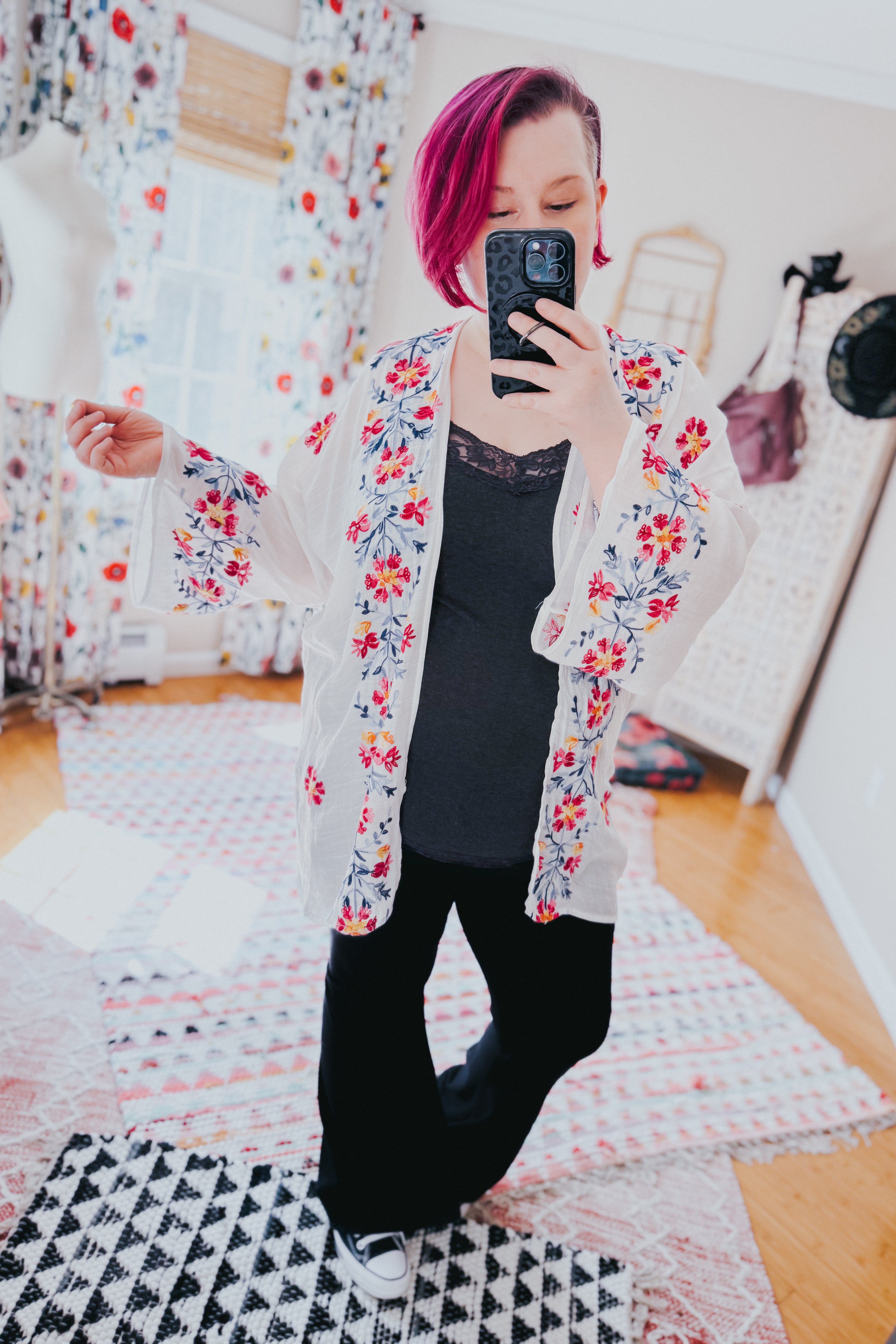 Woman wearing an ivory embroidered duster with colorful floral detailing over a black lace-trim camisole and black pants indoors