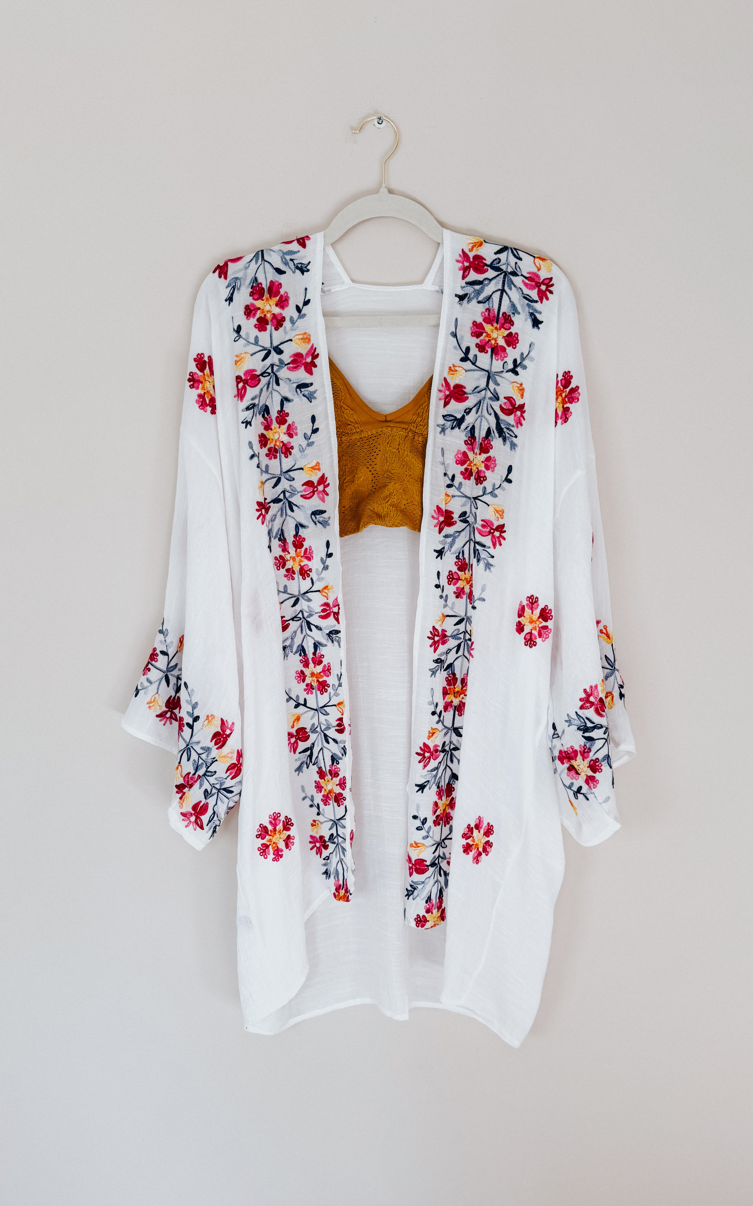 Ivory embroidered duster with colorful floral patterns layered over a mustard crochet top on a hanger against neutral wall