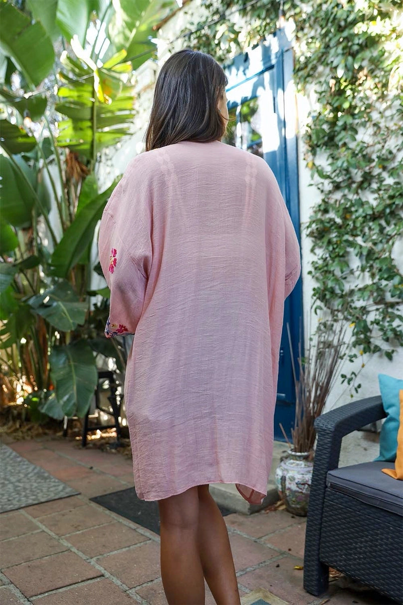Woman wearing a rose-colored embroidered duster standing outdoors near greenery and a blue door in a cozy patio setting
