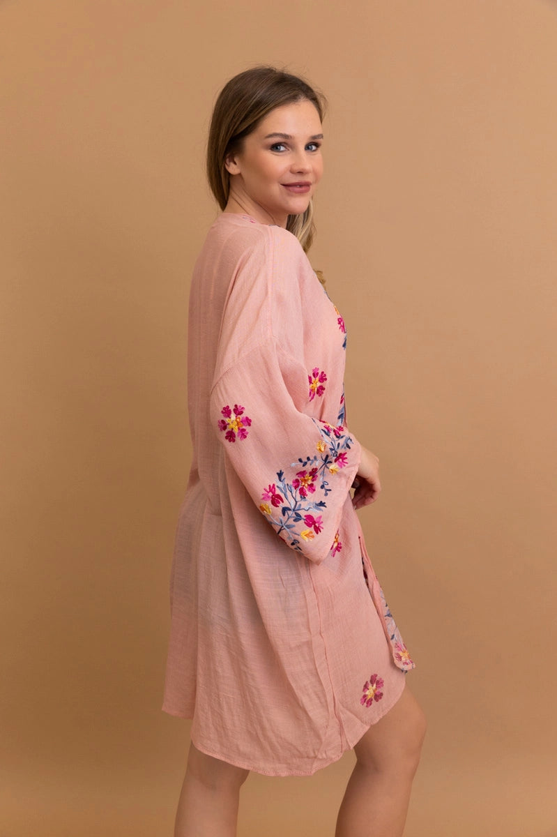 Woman wearing a rose-colored embroidered duster with floral motifs on sleeves and hem, posing sideways against tan backdrop