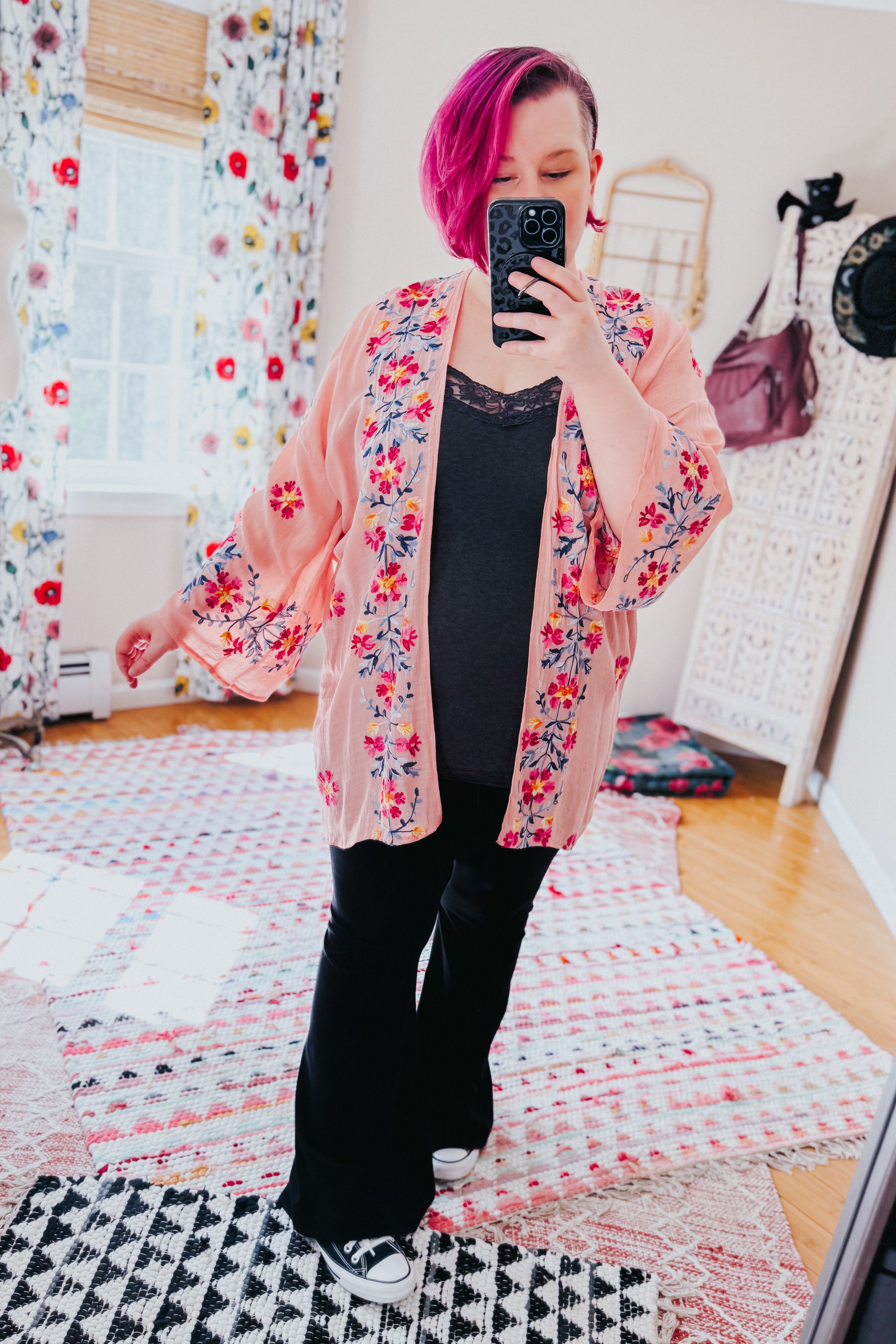Woman with magenta hair wearing a rose embroidered duster over black lace-trim top and black pants in a colorful room