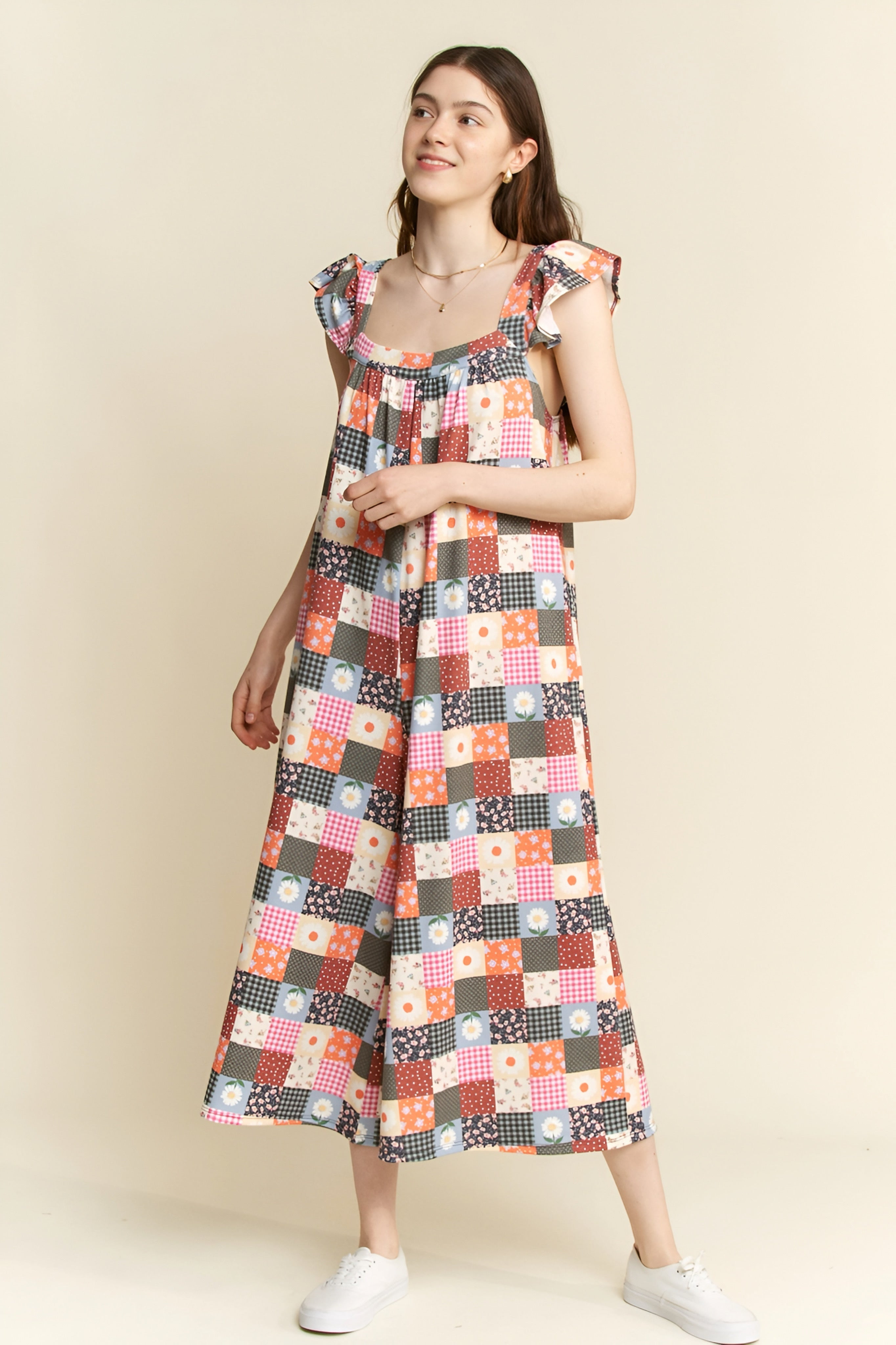 Woman wearing a multicolor patchwork jumpsuit with ruffle sleeves and floral and checkered patterns, standing against beige wall