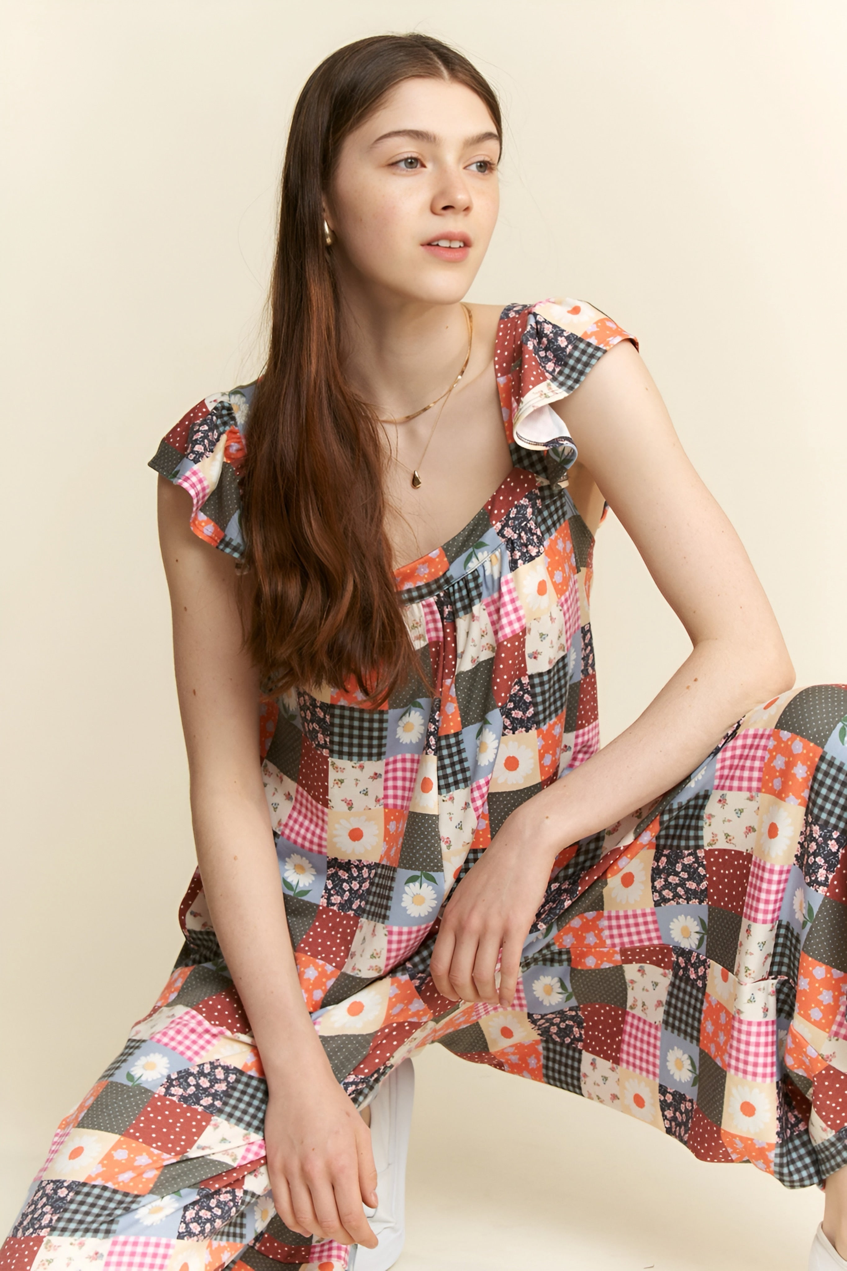 Model wearing a multicolor patchwork jumpsuit with ruffle sleeves, sitting and posing against a beige backdrop