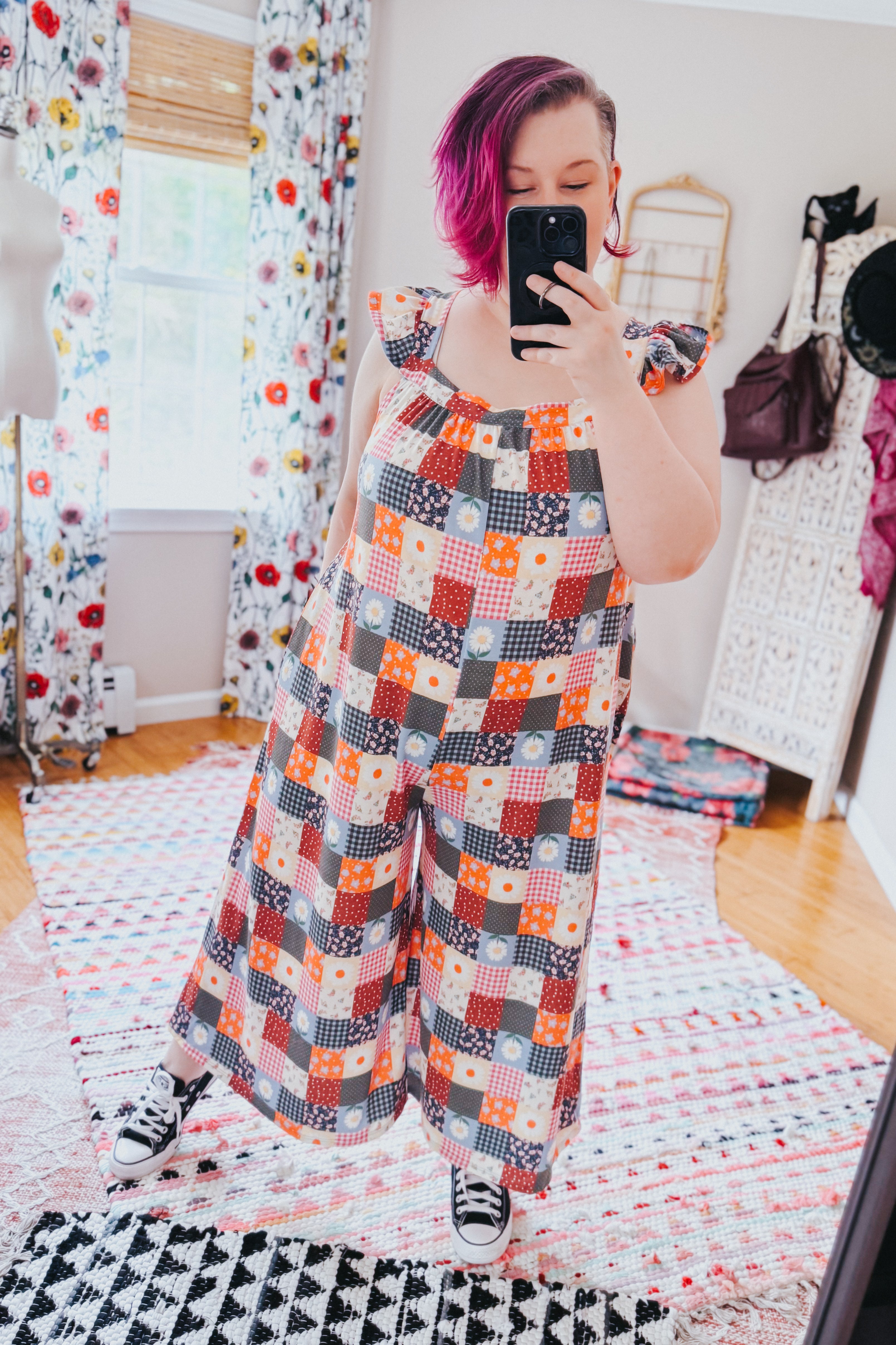 Woman with magenta hair wearing a multicolor patchwork jumpsuit with ruffle sleeves taking a mirror selfie indoors
